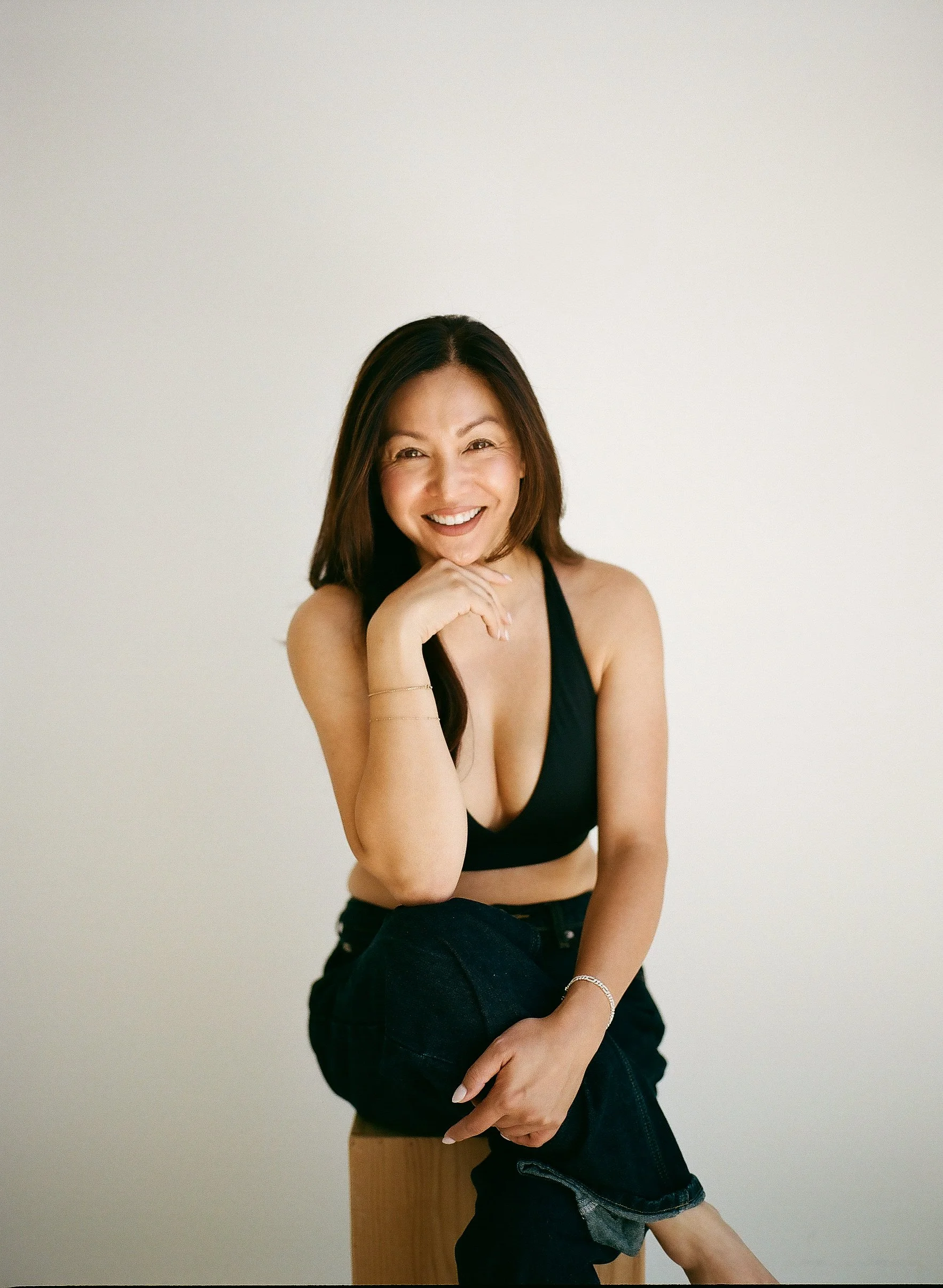A woman wearing a black crop top, denim shorts, and tan tights, smiling and sitting against a plain light-colored wall.