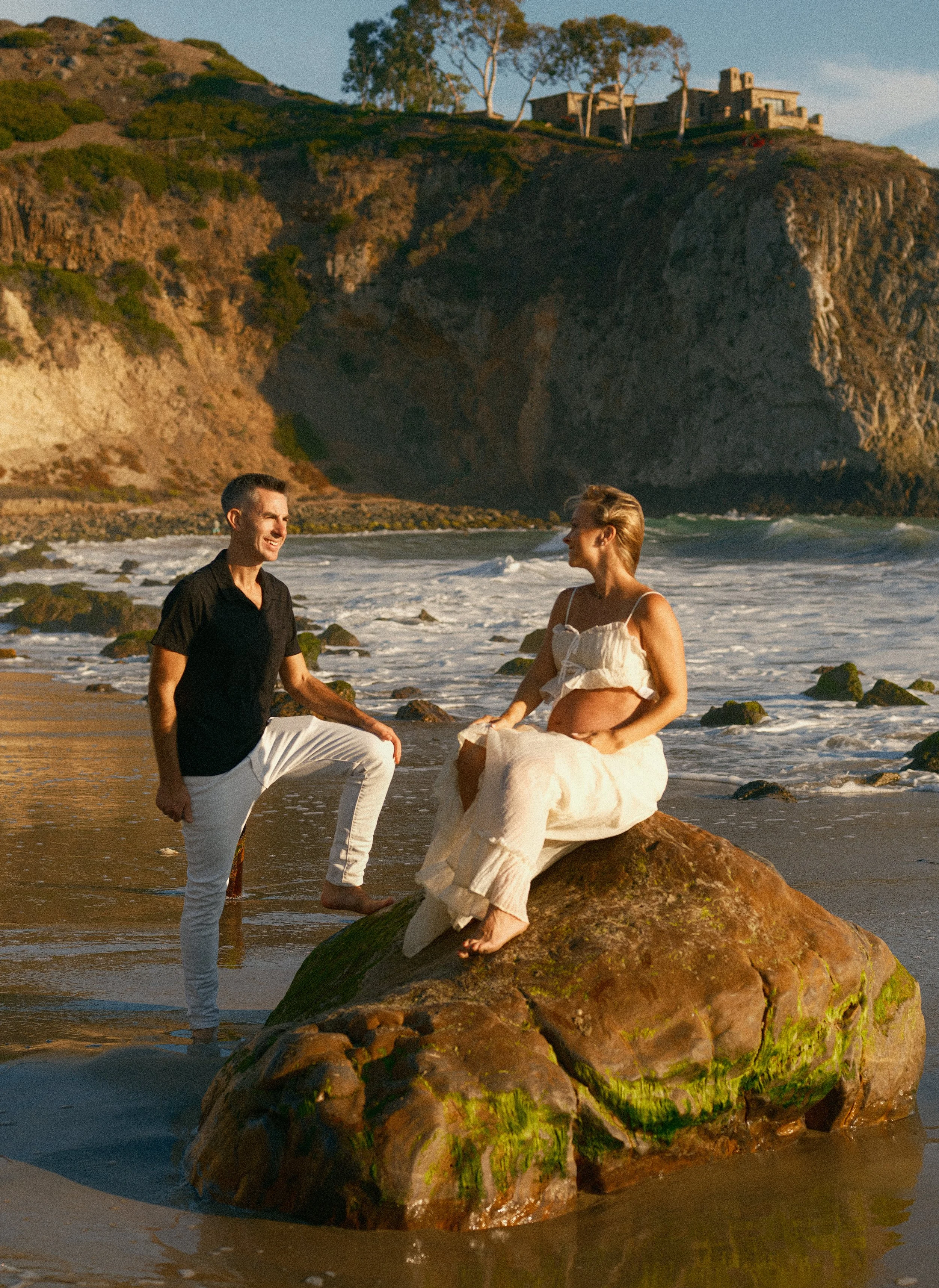 A man and a woman on a beach with rocks, with a large rocky cliff in the background. The man is kneeling on one knee, holding the woman's hand. The woman is sitting on a large rock, pregnant, and wearing a white dress. The scene is during sunset.