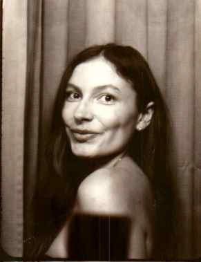 A woman with long dark hair smiling at the camera, sitting in front of curtains, with a dark drink in front of her.