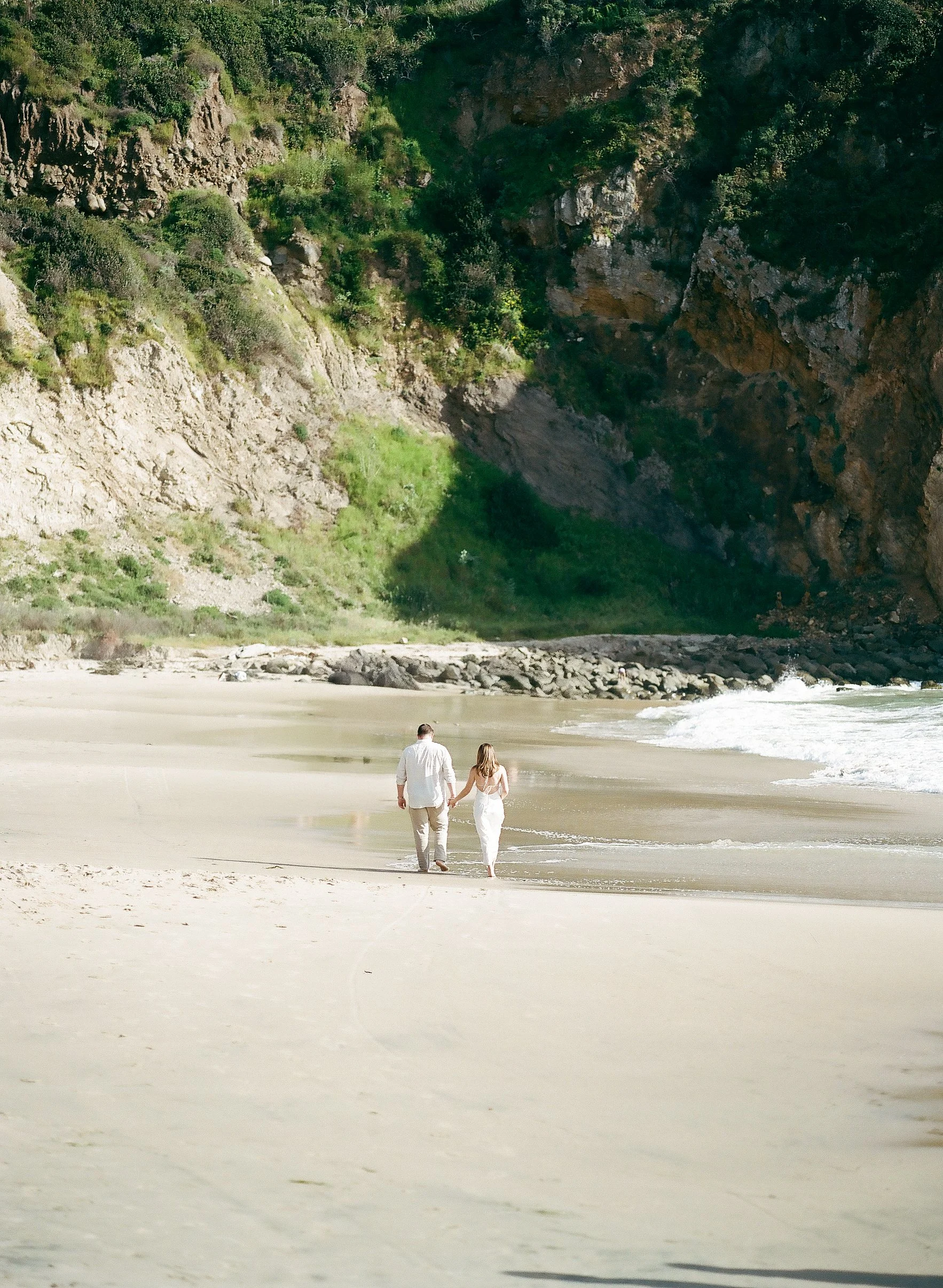 A man in light-colored clothing and a woman in a white dress walking hand in hand along a sandy beach, with cliffs and greenery in the background.