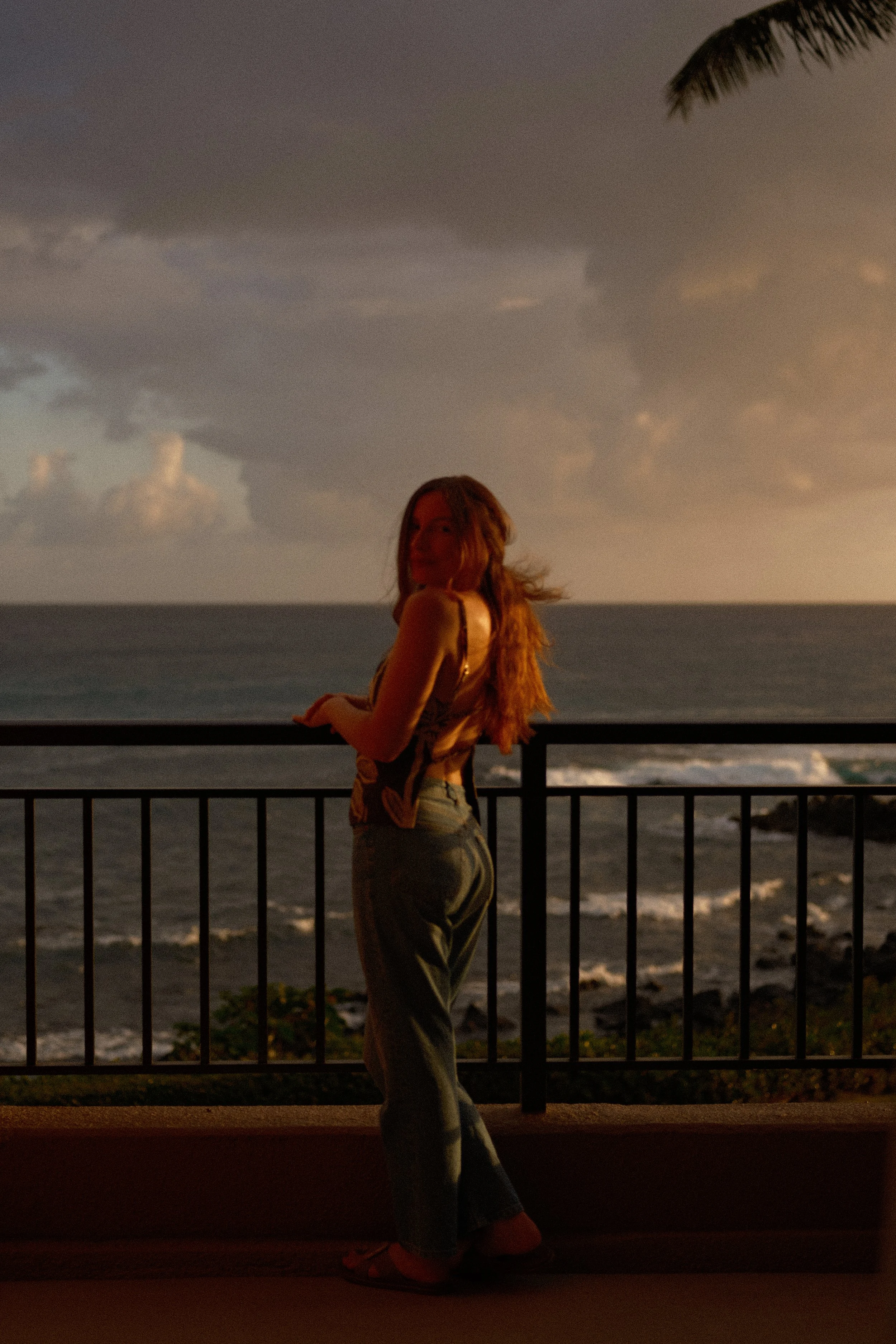A woman with long hair wearing a tank top and pants, standing near a railing on a balcony overlooking the ocean during sunset with dark clouds in the sky.