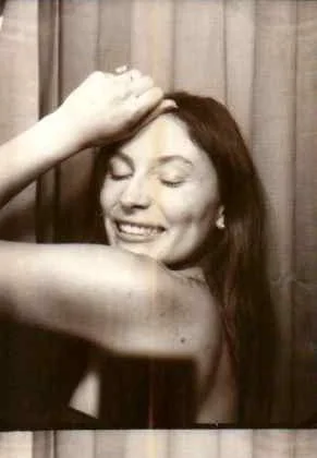 A woman with long hair smiling and resting her hand on her head in front of a curtain.