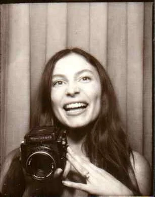 A woman with long, dark hair taking a selfie in front of a curtain, smiling and holding the camera.