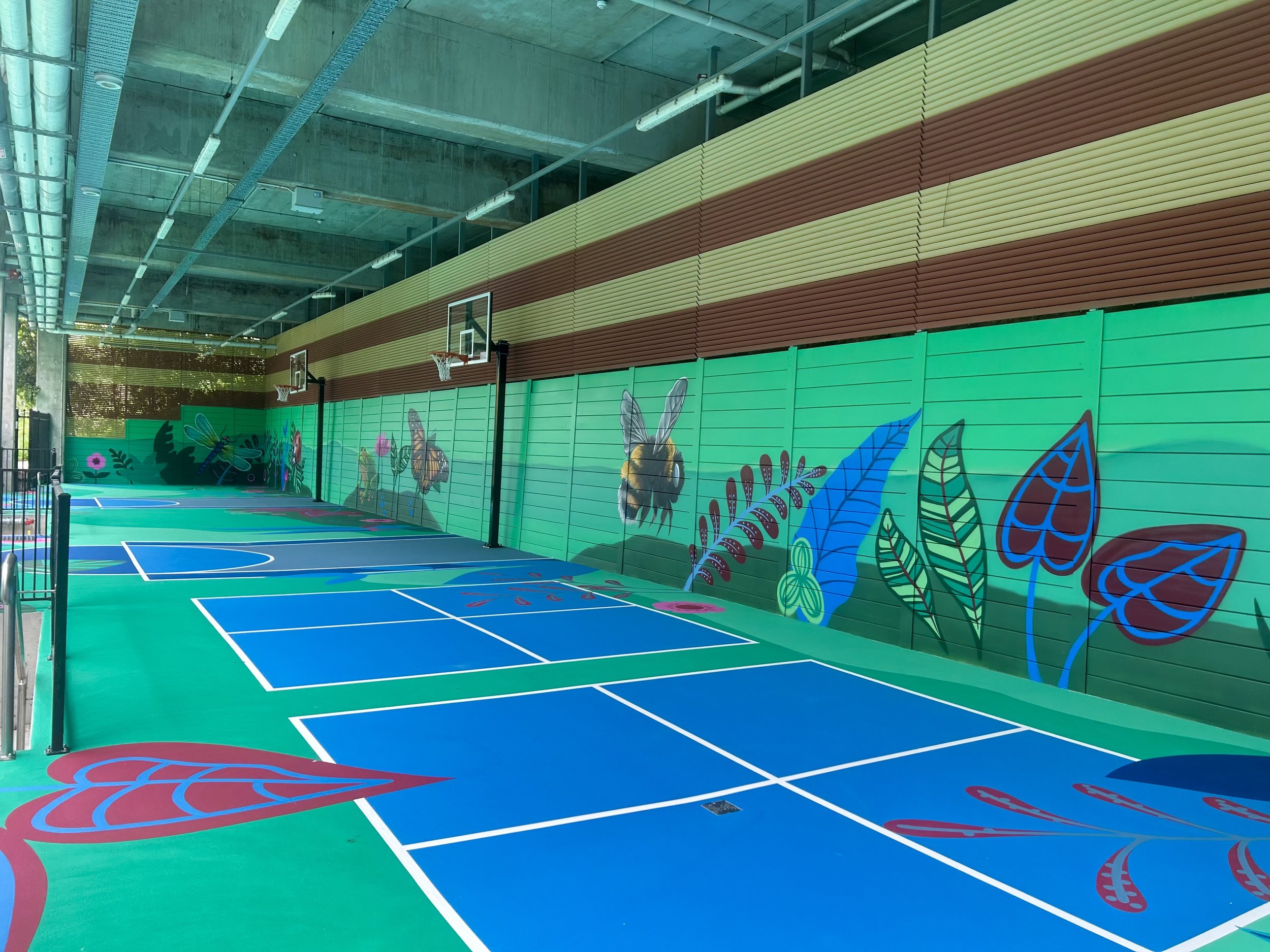 Indoor basketball court with colorful wall mural featuring bees, flowers, and leaves, under a high ceiling with exposed pipes and lighting.