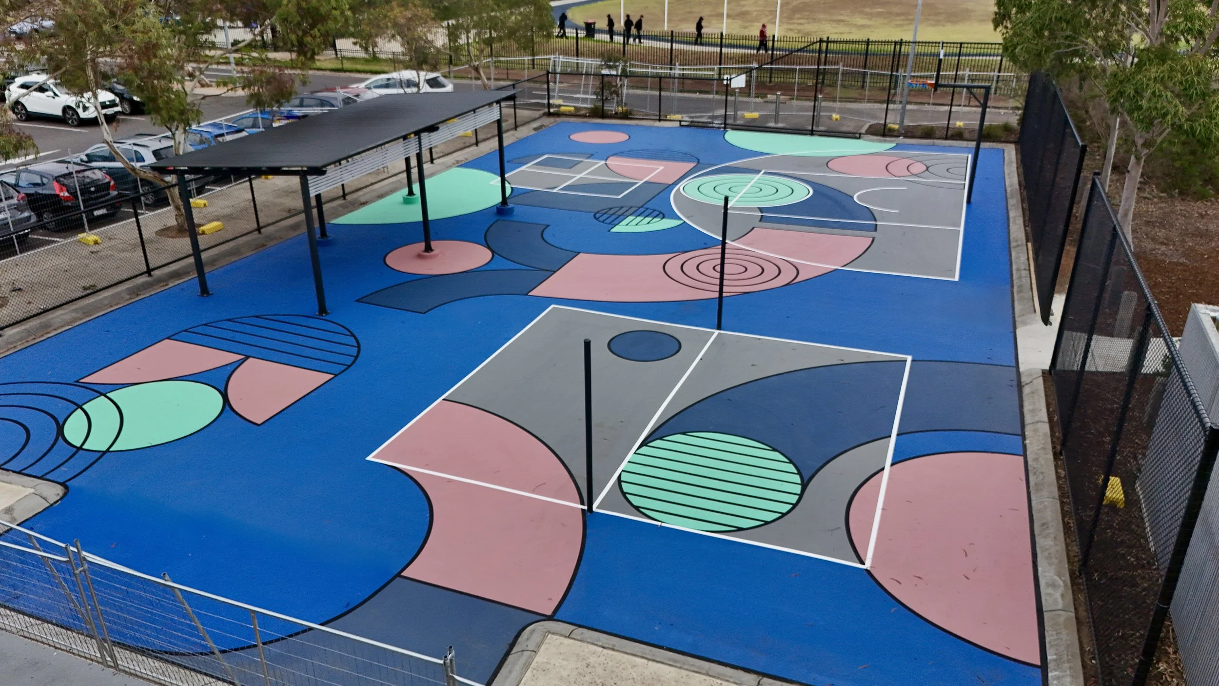 Colorful outdoor basketball court with various geometric shapes and patterns, surrounded by a black fence, with a parking lot and trees in the background.