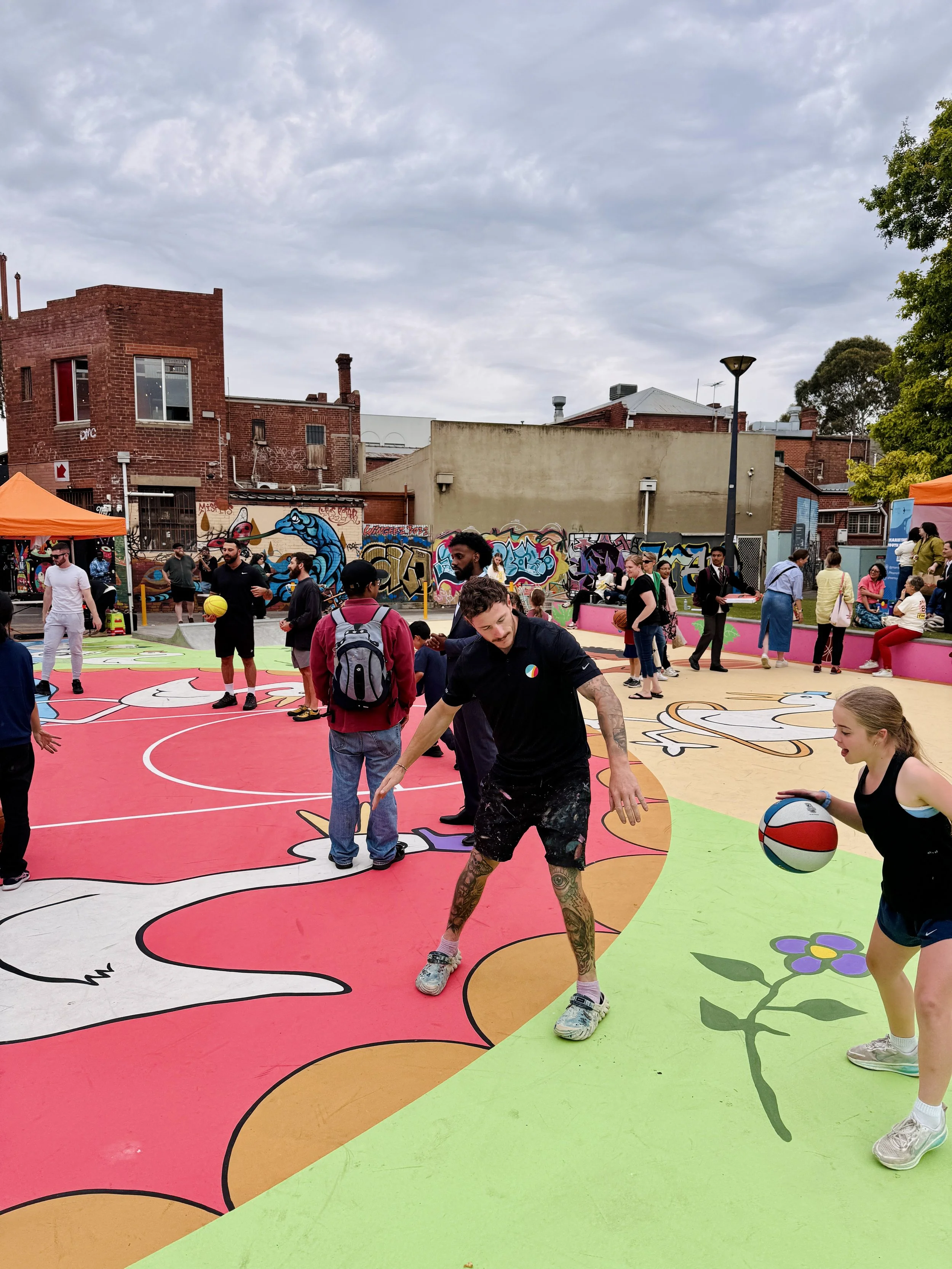 People playing basketball on a colorful outdoor court with graffiti walls and a cloudy sky.