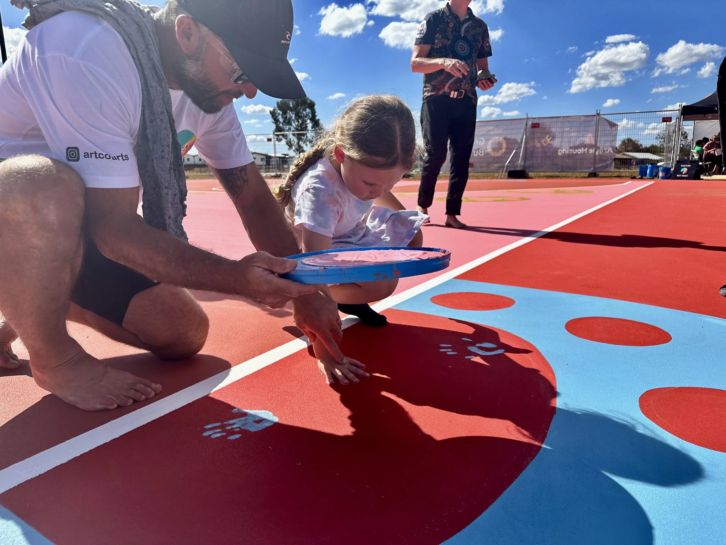 A man and a young girl are playing on a colorful outdoor court with pink, blue, and red markings. The man is kneeling and painting paw prints on the ground, while the girl is sitting and watching. Other people are visible in the background under sunny skies.