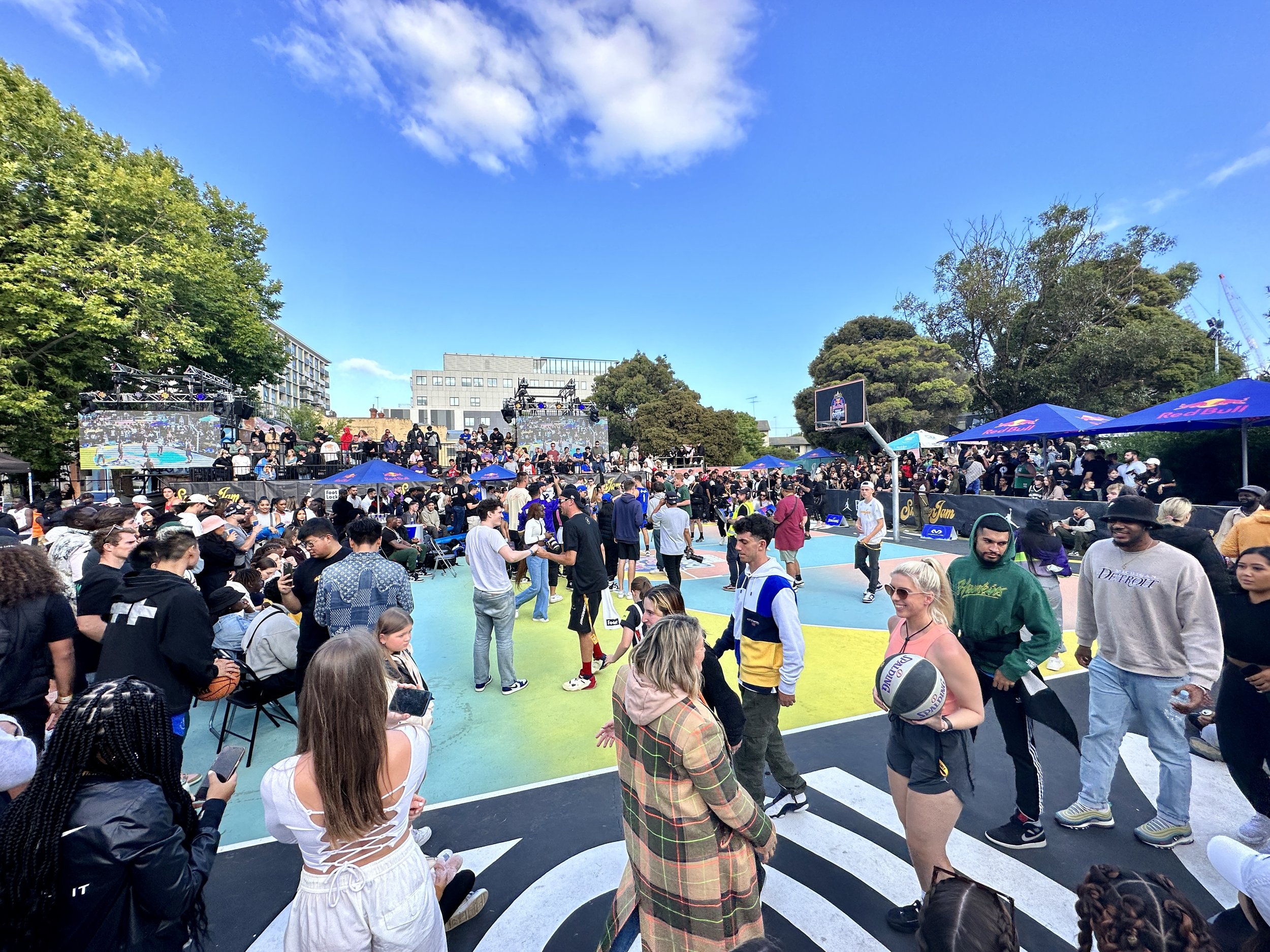 Crowd gathered at an outdoor basketball court during a sunny day, with large LED screens, trees, and buildings in the background.