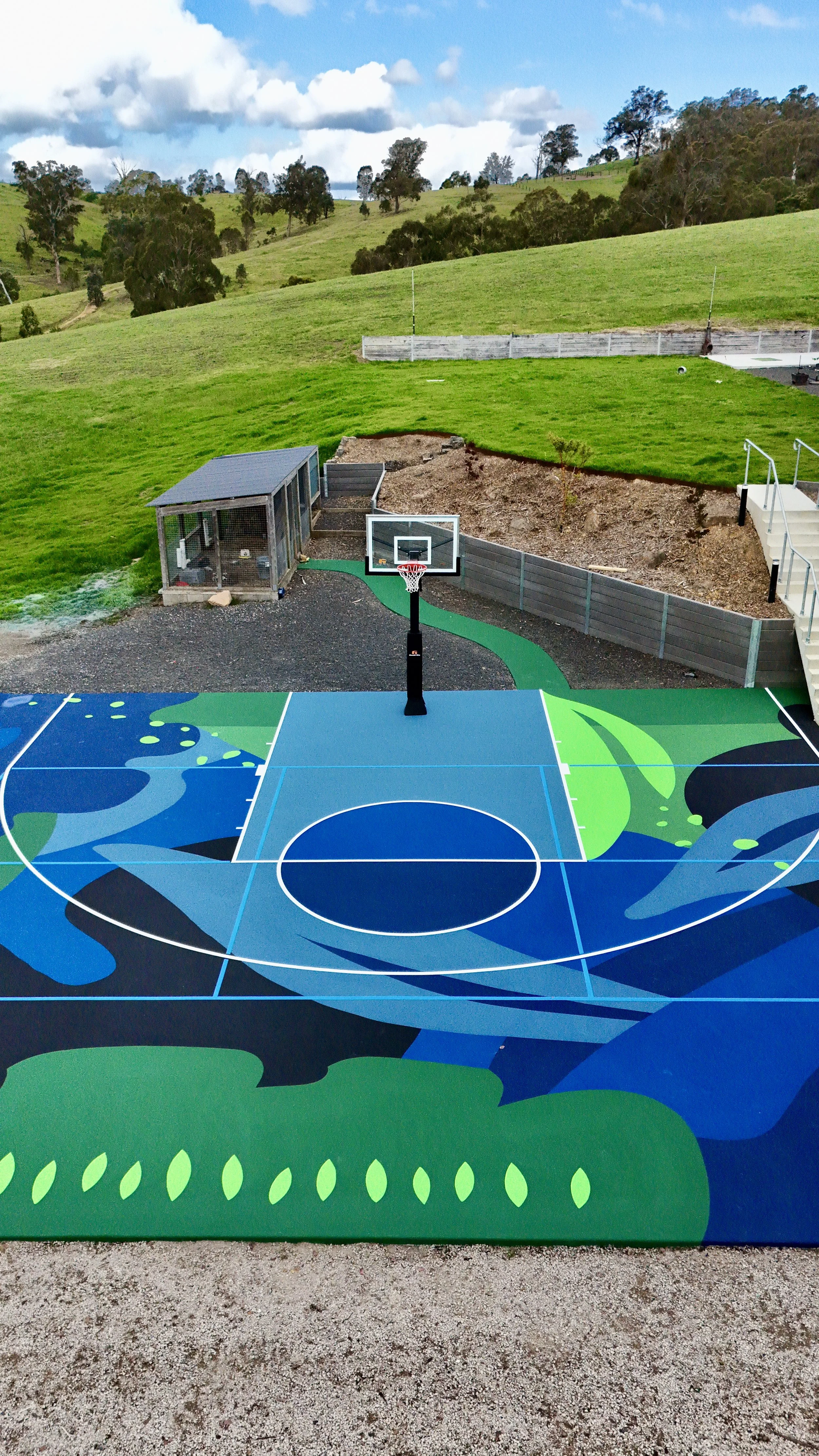 Colorful outdoor basketball court with a mural design, a hoop, and a small fenced area next to a grassy hillside and distant trees under a partly cloudy sky.