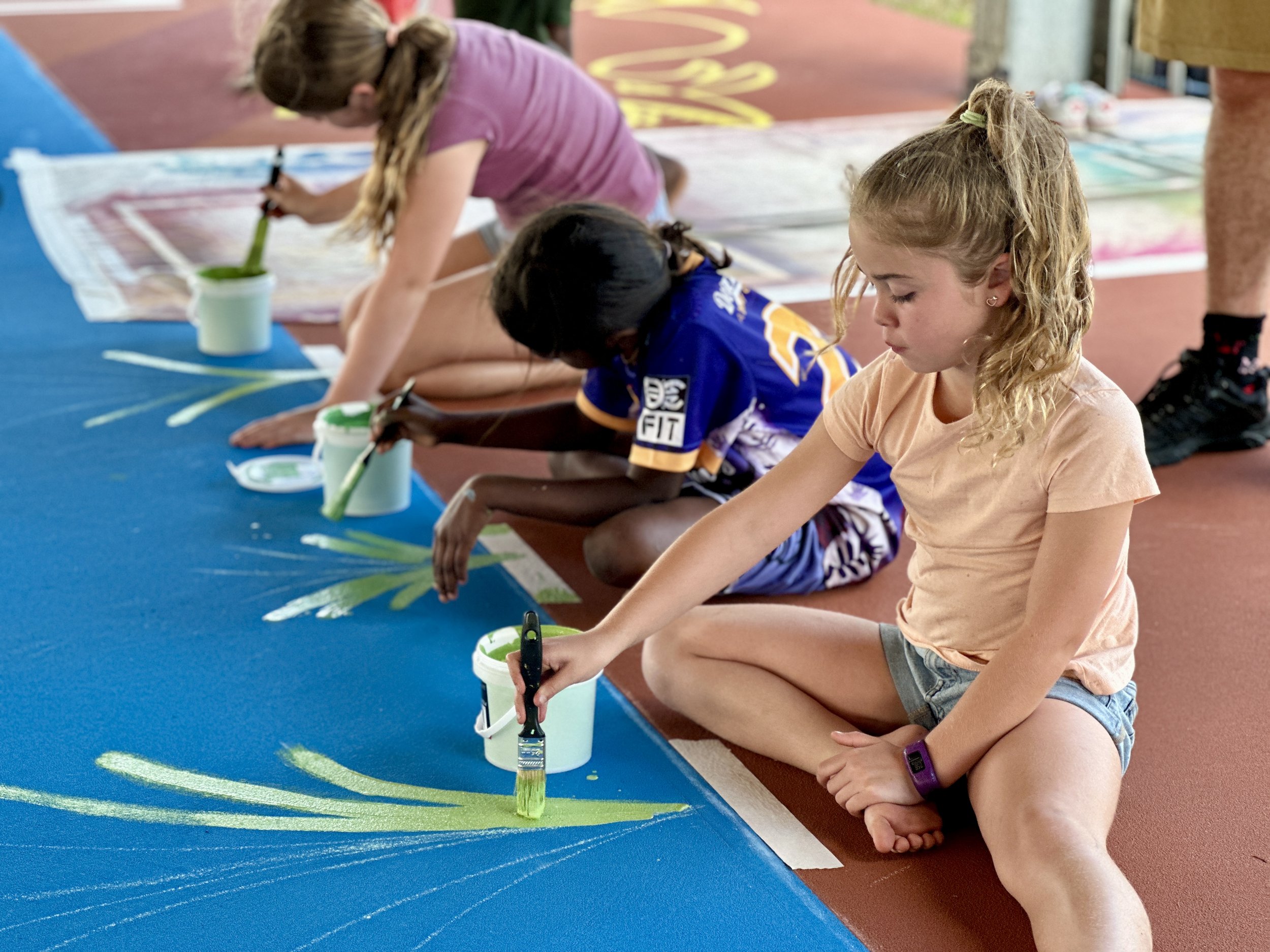 Children are kneeling on the floor, painting a colorful design on a large blue surface with brushes and white containers of green paint.