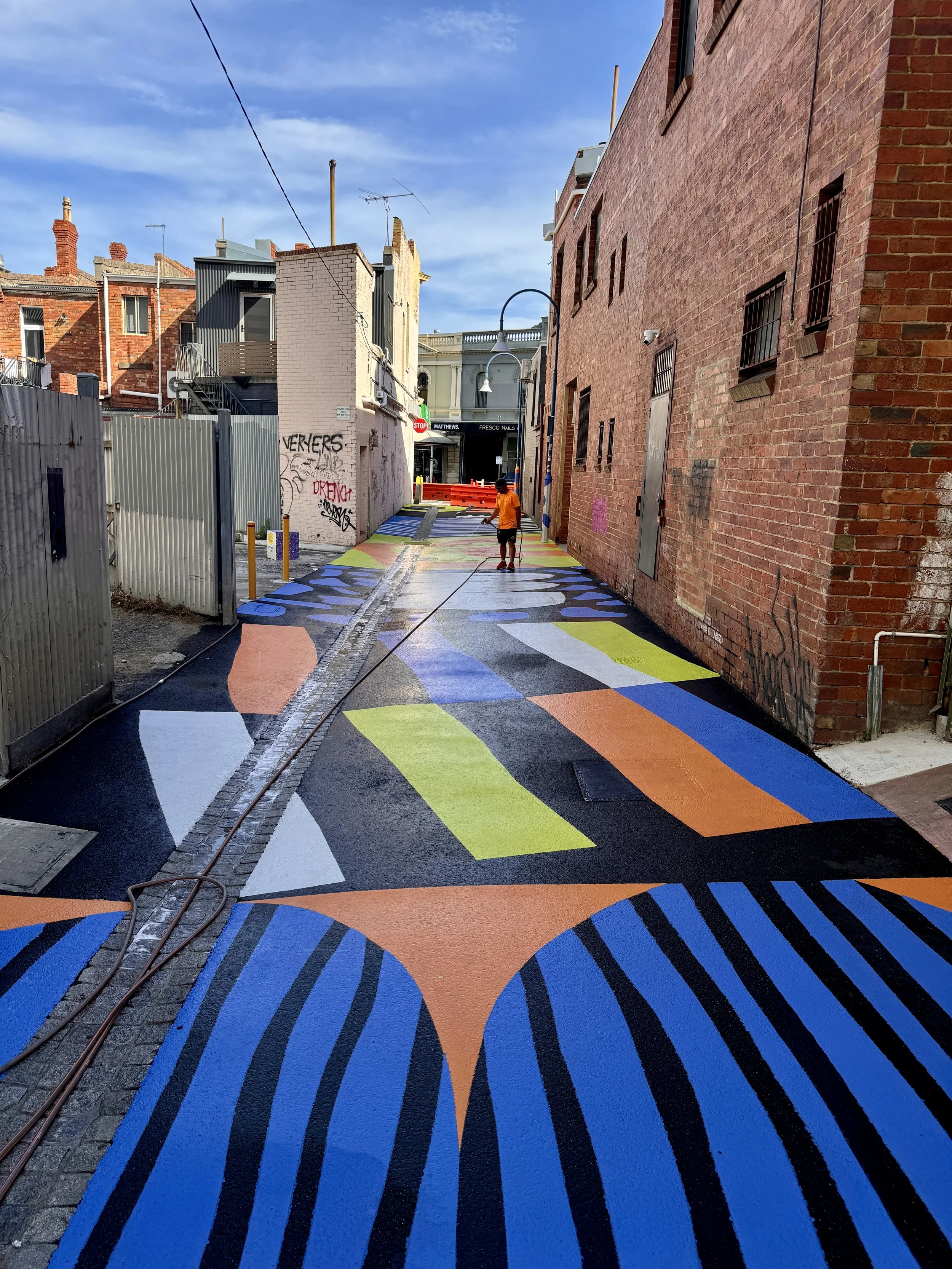 City alleyway with colorful, geometric street art painted on the pavement, including patterns in blue, orange, green, and white, with brick buildings on either side and a person in an orange shirt painting the street.