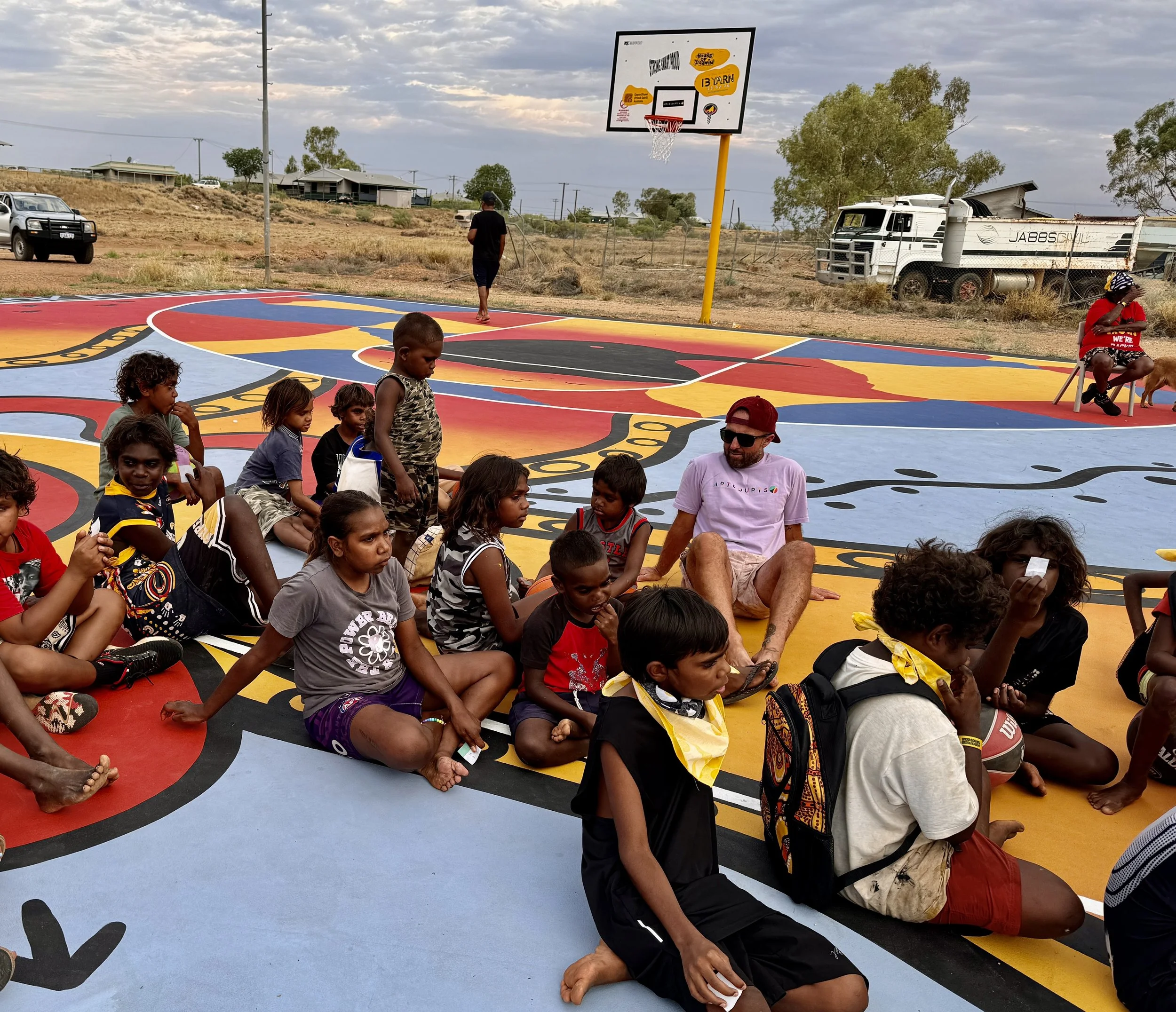 Children and adults gathered on a colorful painted outdoor basketball court in a rural area, with some children sitting and others standing. A man with glasses and a beard is sitting in the foreground. A person wearing a red shirt, black hat, and patterned shorts is sitting on a chair with a dog nearby. In the background, there is a basketball hoop, a white truck with equipment on the side, parked vehicles, trees, houses, and an overcast sky.