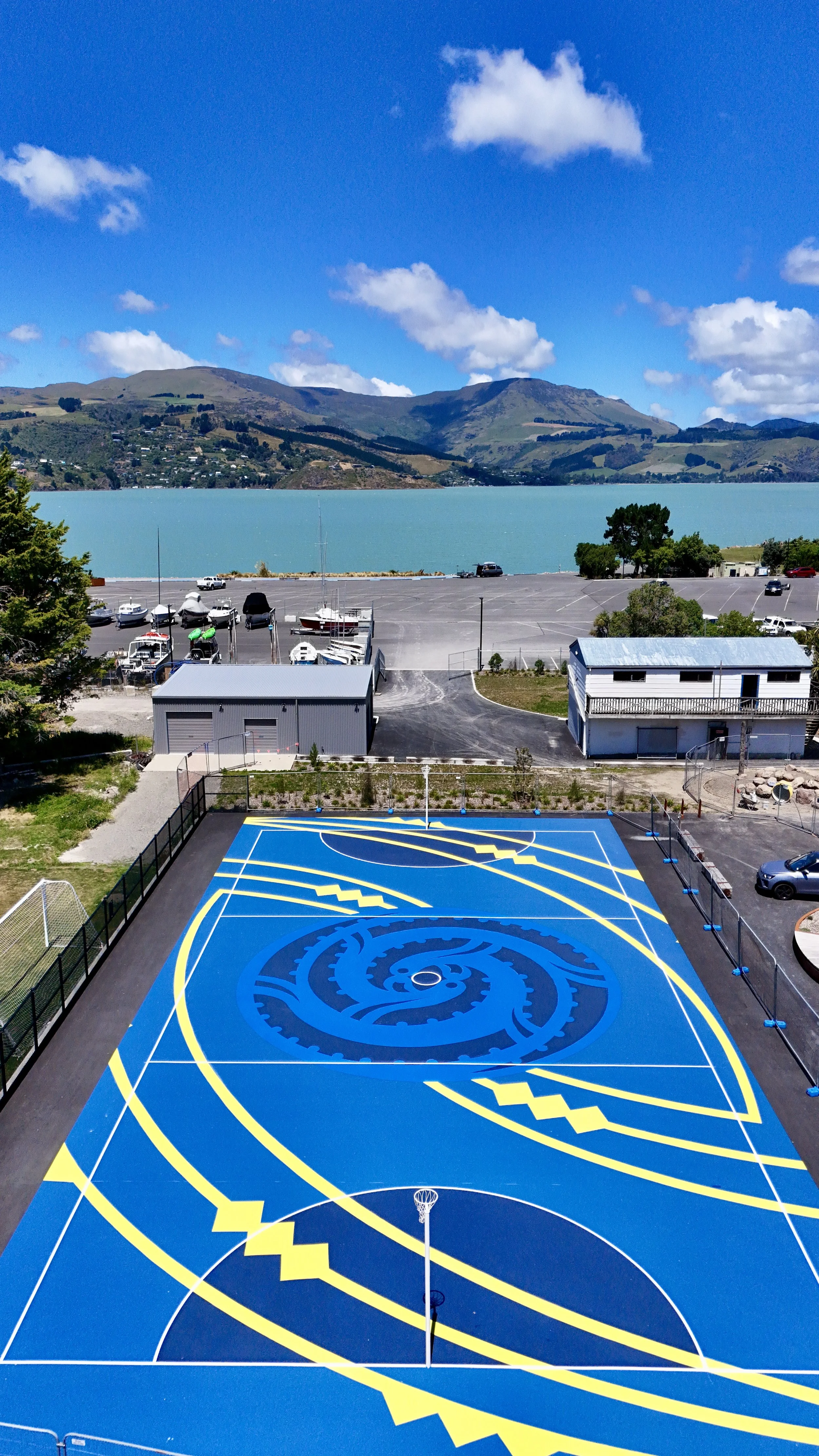 Colorful basketball court with a spiral and zigzag design in blue, yellow, and black, located outdoors near a parking lot, with a lake and mountains in the background under a partly cloudy sky.