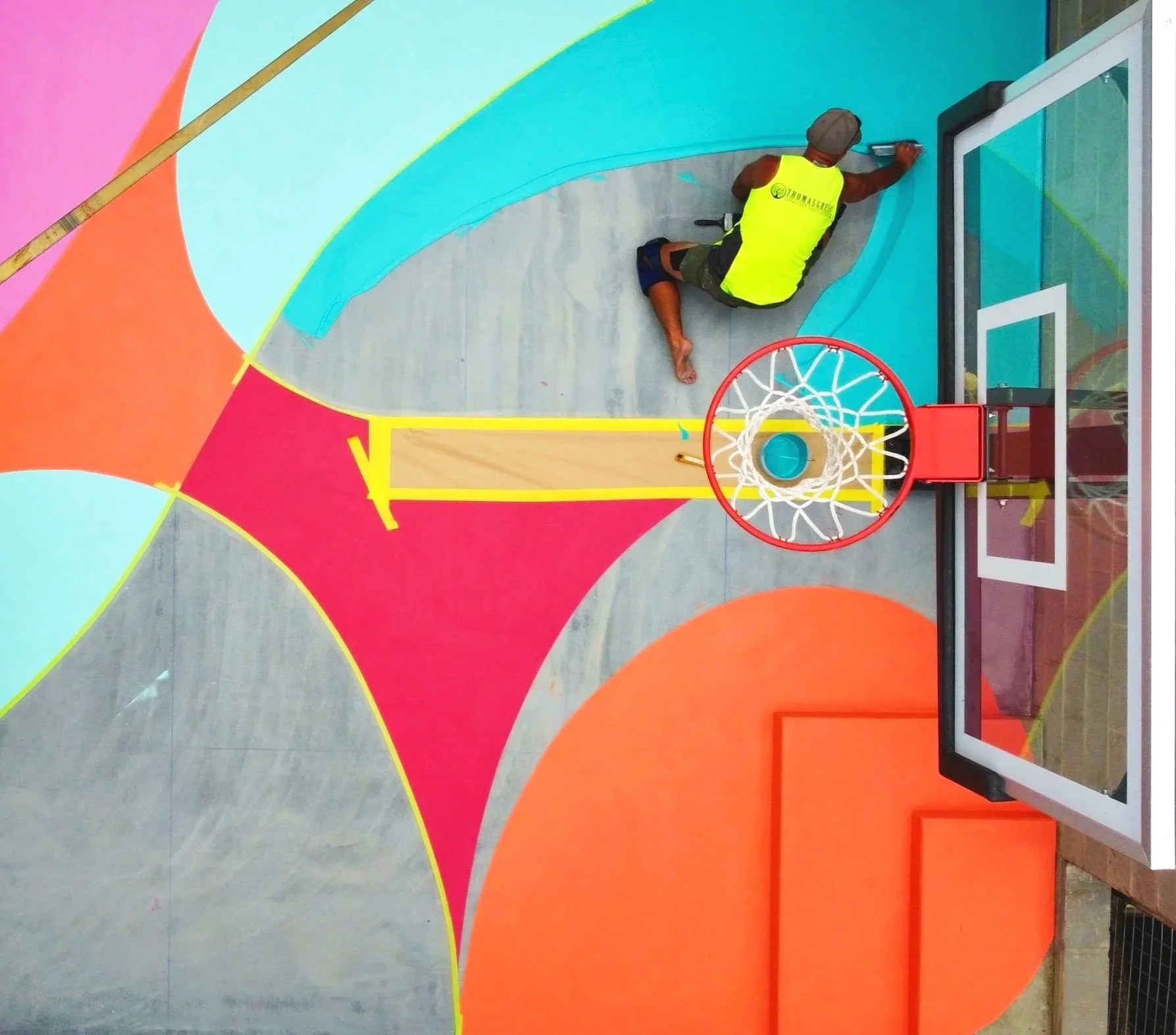 A man painting a colorful mural on a wall, which includes a basketball court with a hoop, in an indoor gymnasium.