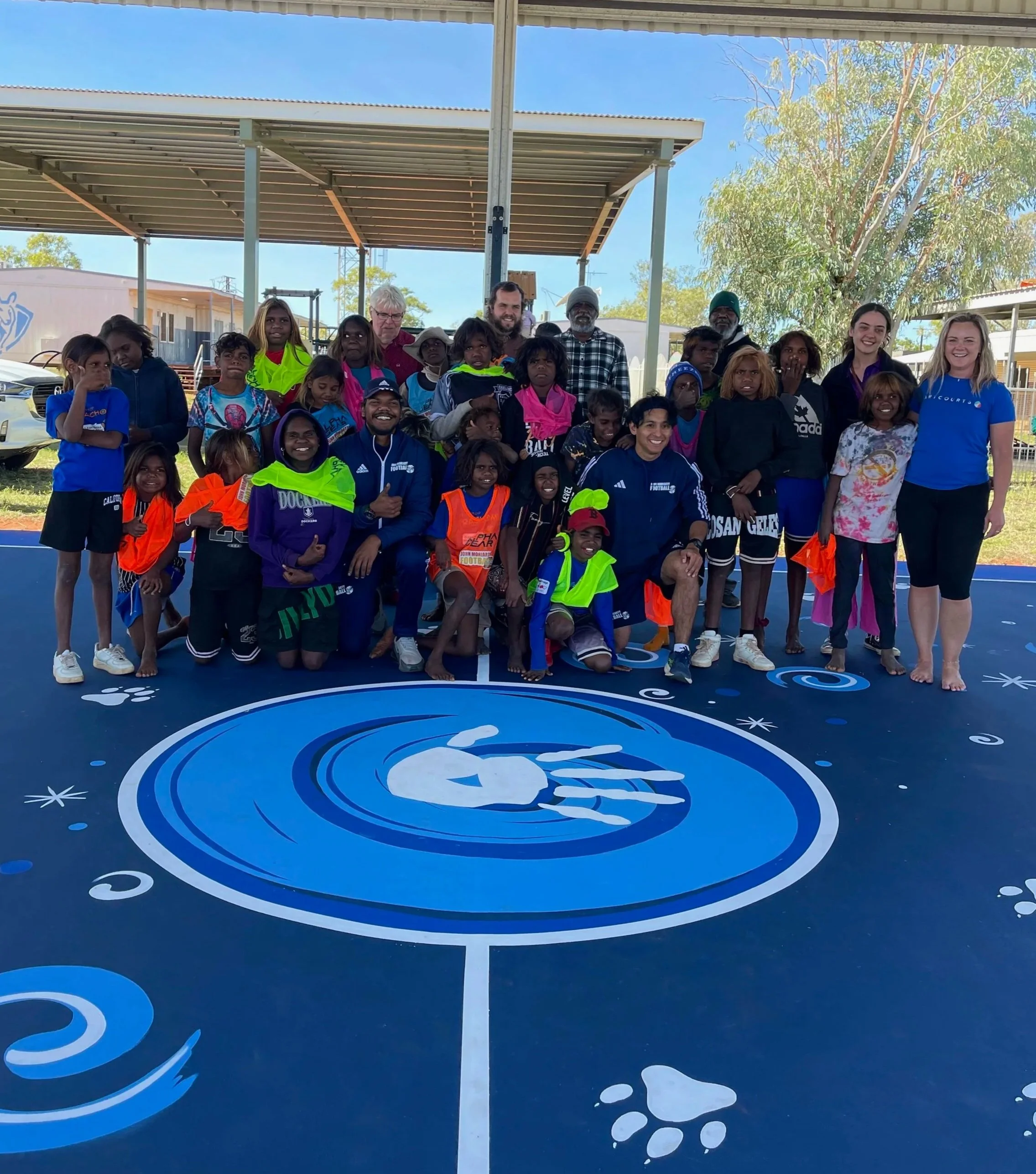 Group of children and adults posing for a photo on a decorated outdoor basketball court with a space-themed design, under a shaded structure with trees and buildings in the background.