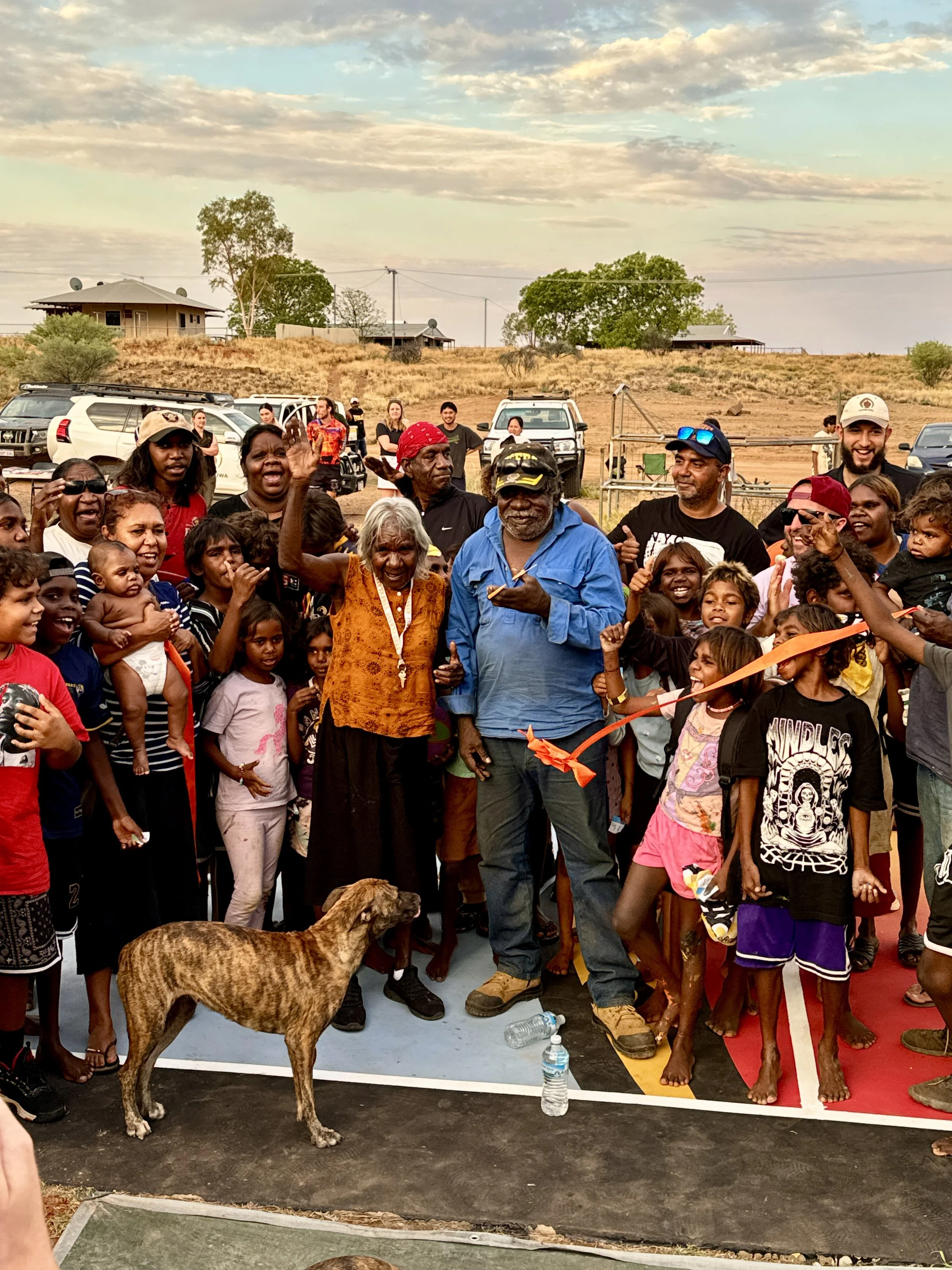 A large group of people, including children and adults, standing together outdoors on a dry field with some houses and trees in the background, and a few cars parked behind them. There is a dog in the foreground, and some people are smiling and raising their hands. The sky is partly cloudy.