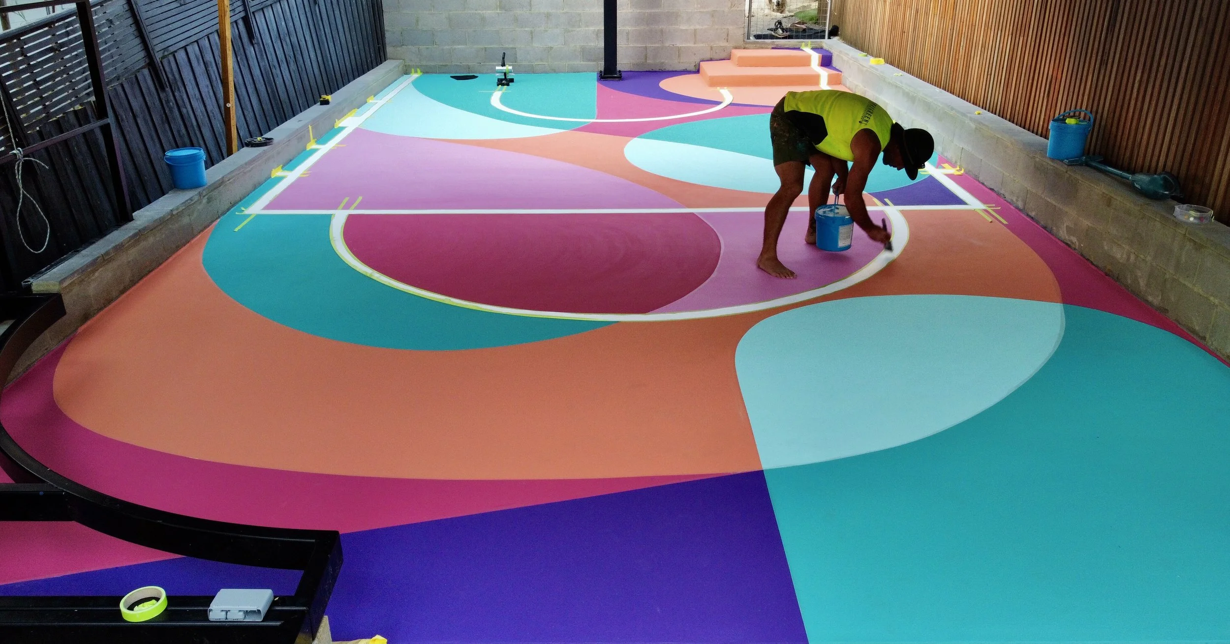 A person painting a colorful abstract mural on a floor with various shapes and colors including pink, blue, teal, orange, and purple, using a brush and paint bucket.