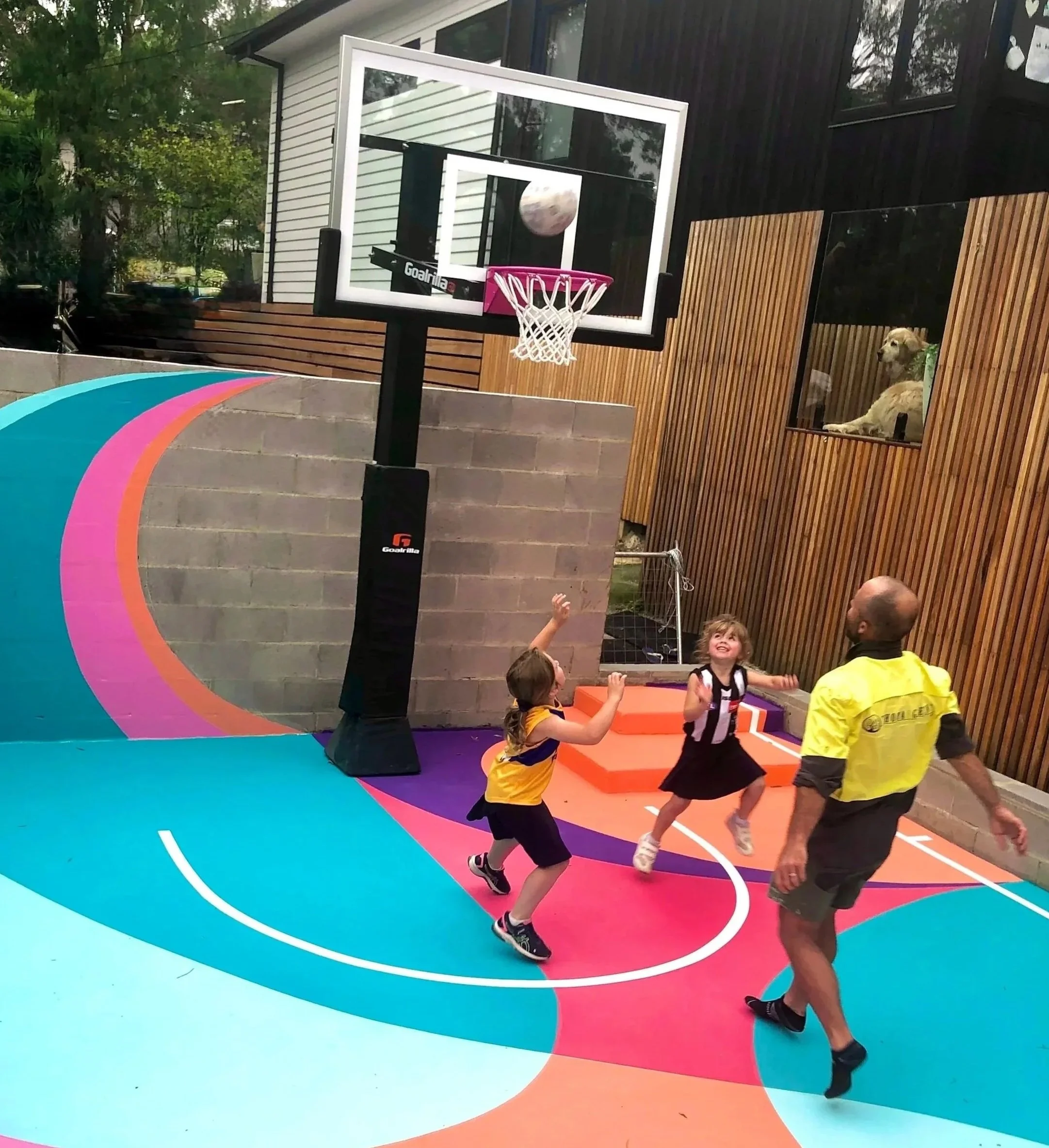Children playing basketball on a colorful court while a man in a yellow shirt watches. A dog is sitting behind a wooden fence, observing the scene.