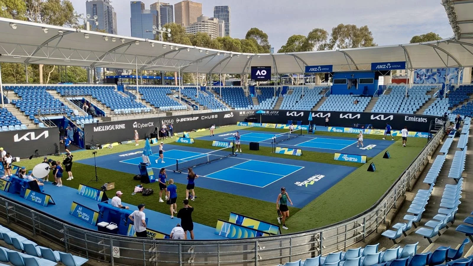 An outdoor tennis court with players practicing during a tournament, surrounded by empty blue seats and stadium seating, with city buildings in the background.