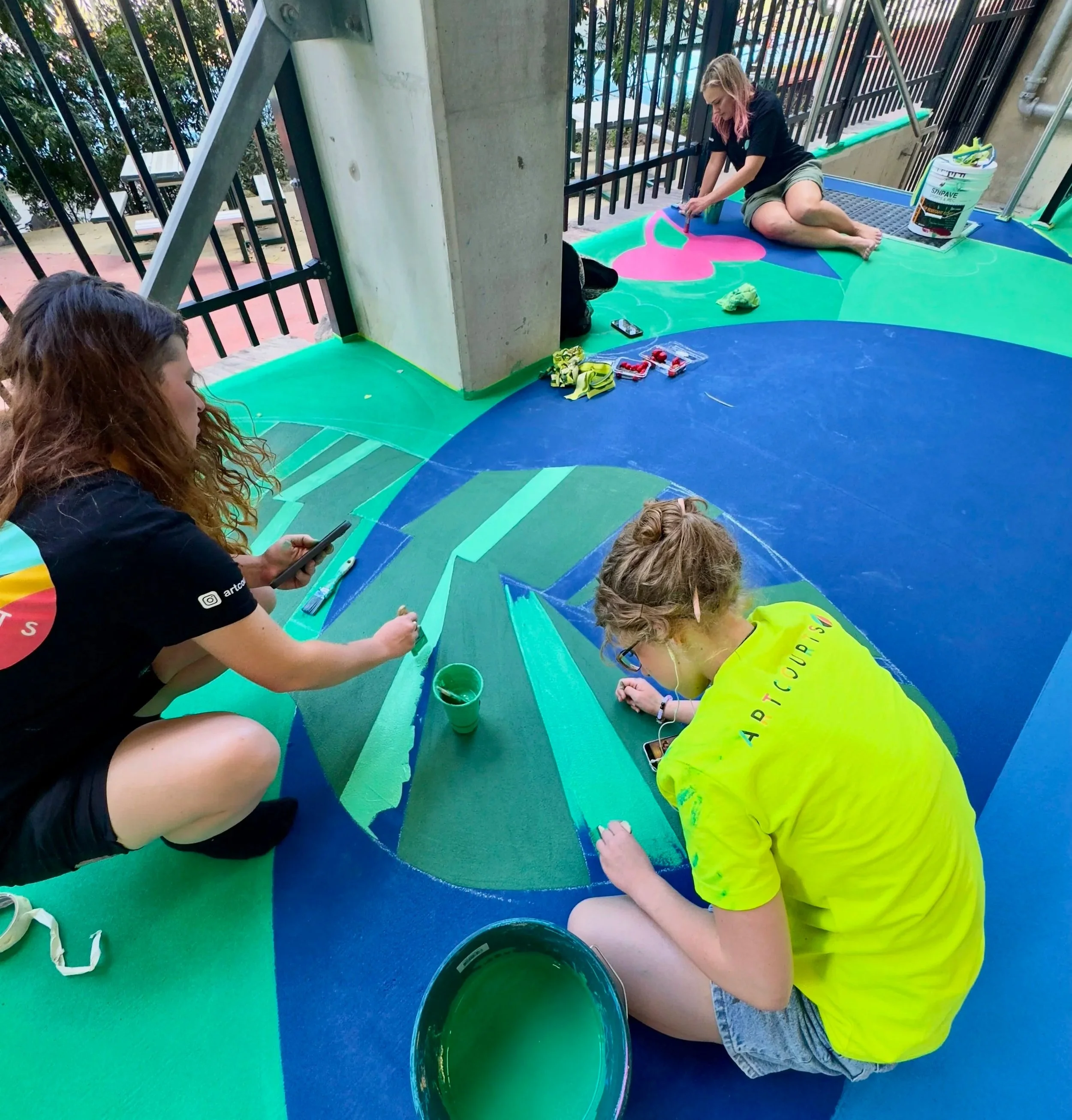 Several people crouch on a colorful floor, painting a mural with bright colors. They are using paint and brushes, and there are containers of paint around them.