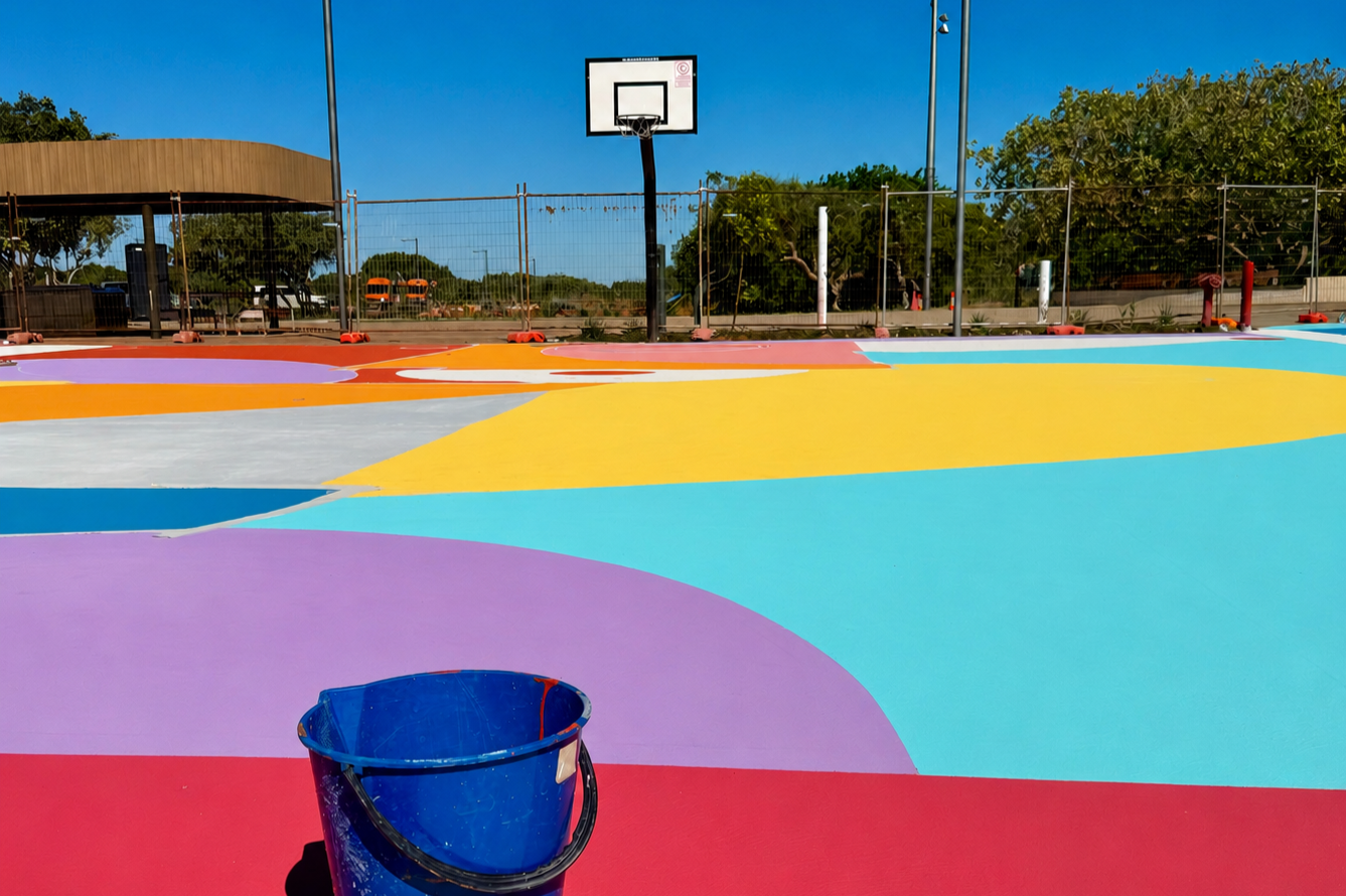 A colorful outdoor basketball court with painted geometric shapes, a basketball hoop, and a blue bucket in the foreground, under a clear blue sky.
