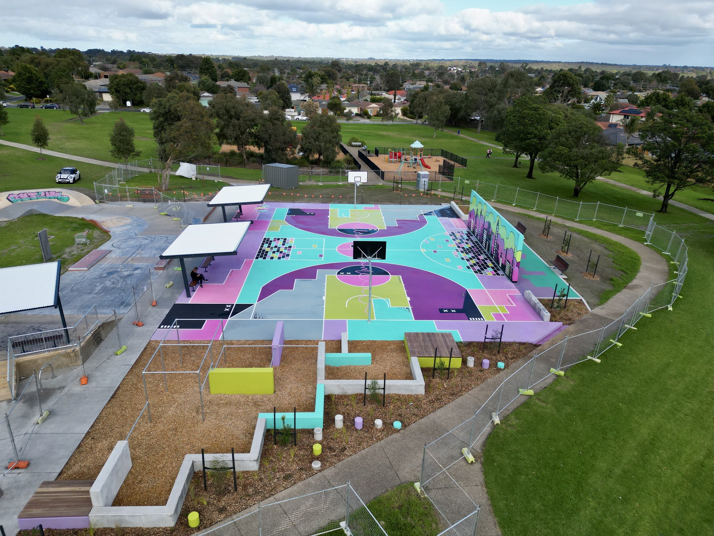 A colorful outdoor basketball court with painted lines and designs, surrounded by green grass, trees, and a neighborhood with houses, and a playground in the background.