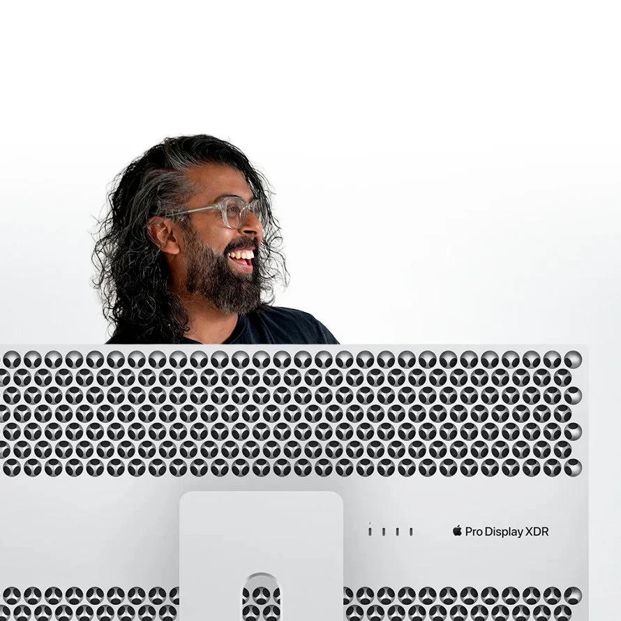 A man with long curly hair and a beard smiling and looking to his left, sitting behind an Apple Pro Display XDR monitor with a white background.