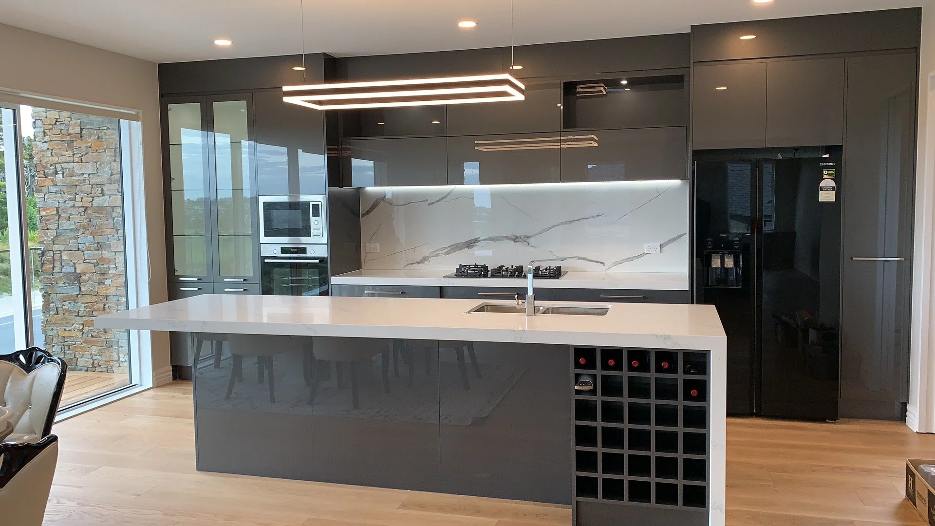 Modern kitchen with black cabinetry, white marble backsplash, and a white kitchen island with a built-in sink. There's a large black refrigerator and a microwave oven. The cabinetry has glass doors, and a geometric light fixture hangs from the ceilin