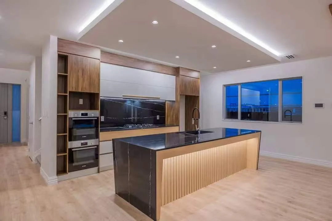 Modern kitchen with a black marble island, wooden cabinets, built-in ovens, and large window showing a blue evening sky.