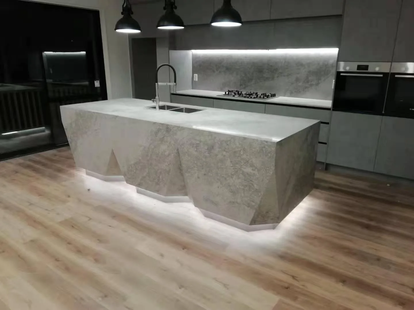 Modern kitchen with a large, angular stone island with an under-light glow, a built-in sink, black faucet, gray cabinetry, stainless steel oven and microwave, and a wall-mounted gas stove, illuminated by black pendant lights.