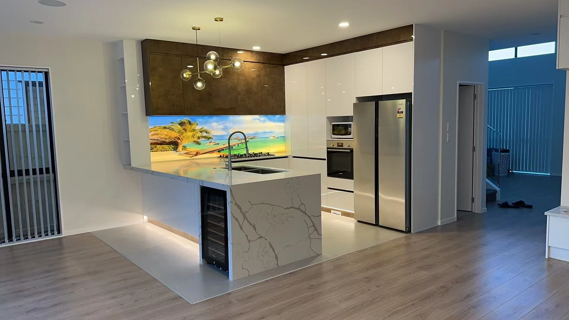 Modern kitchen with a marble island, white and dark brown cabinetry, stainless steel appliances, a tropical beach mural backsplash, and wooden flooring.