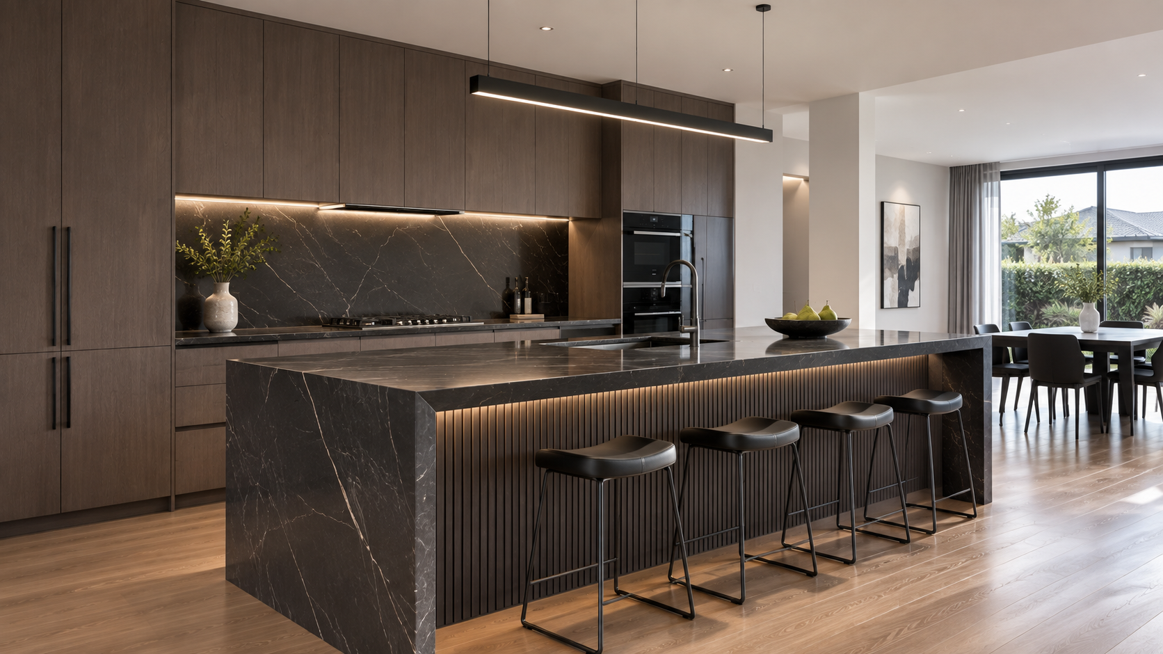 Modern kitchen with black marble island, wooden cabinets, and black bar stools, adjacent to a dining area with a table and chairs, large window with curtains, and outside greenery.