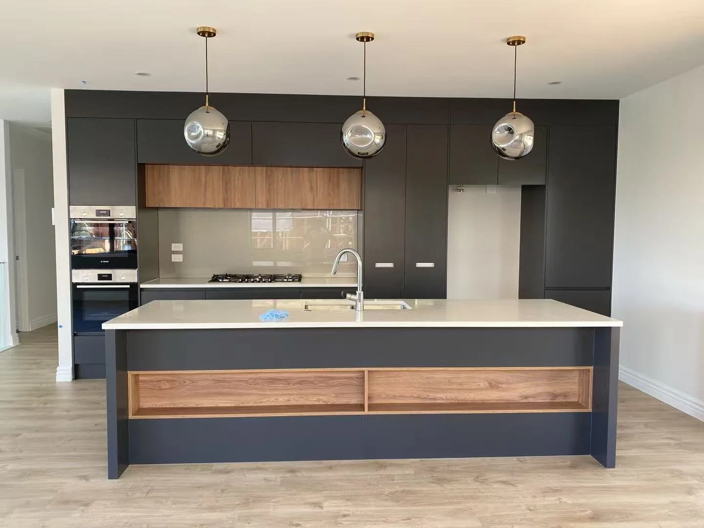 Modern kitchen with dark cabinetry, a white countertop island with wooden accents, three spherical hanging pendant lights, built-in oven and microwave, stovetop, and a sink.