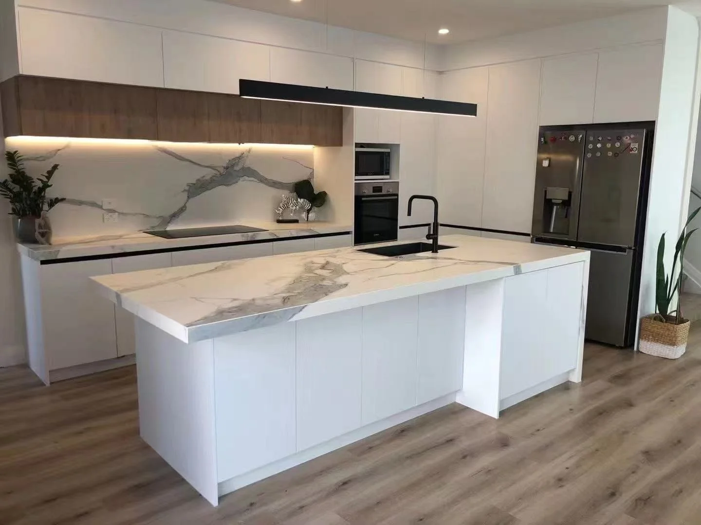 Modern kitchen with white cabinetry, marble countertops, black faucet, built-in oven, microwave, and stainless steel refrigerator. Potted plants and decorative items on the counters, with wood flooring.