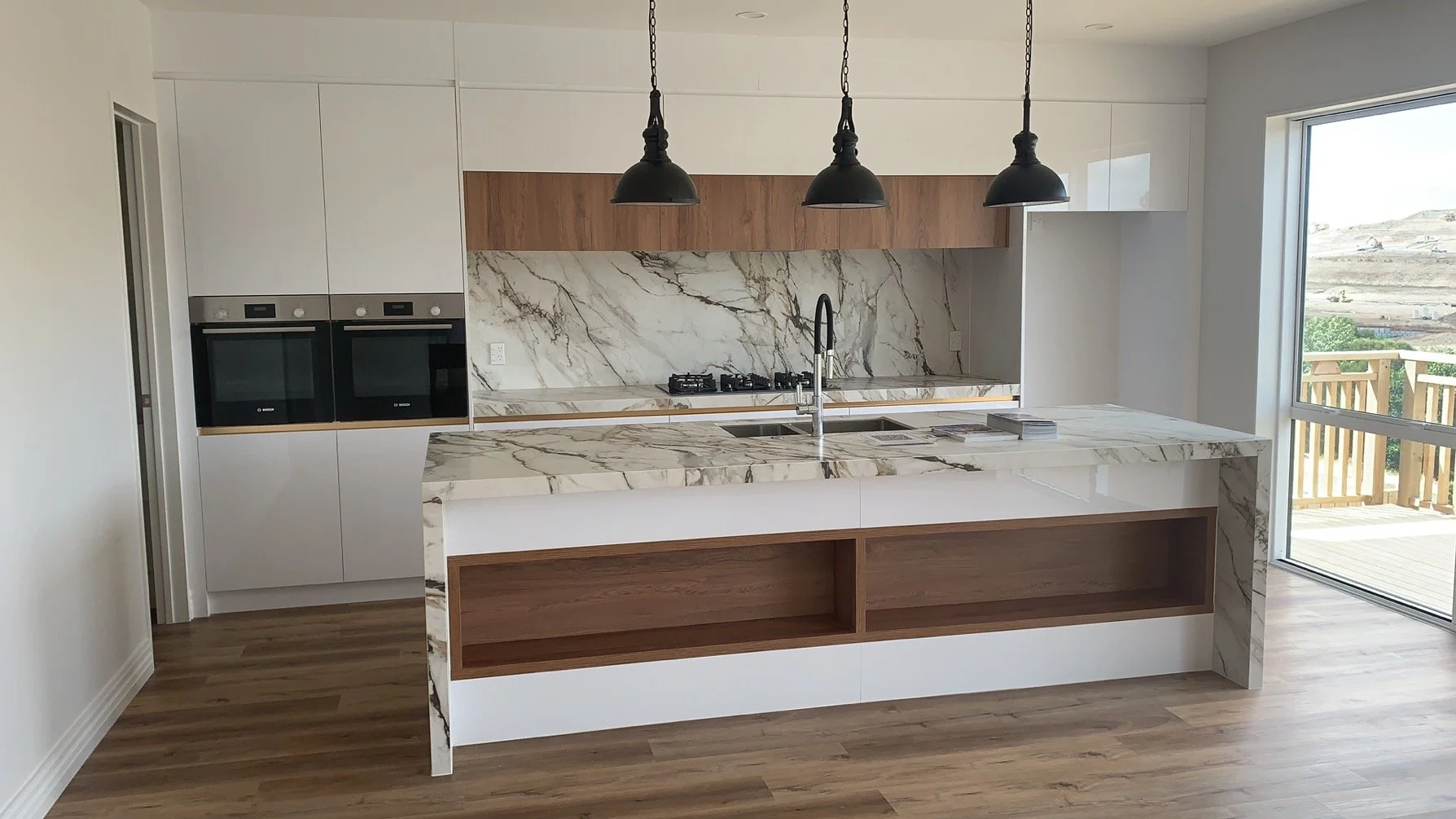 Modern kitchen with a marble island, built-in ovens, a stovetop, a stainless steel sink, and black pendant lights, with a sliding glass door leading to an outdoor deck.