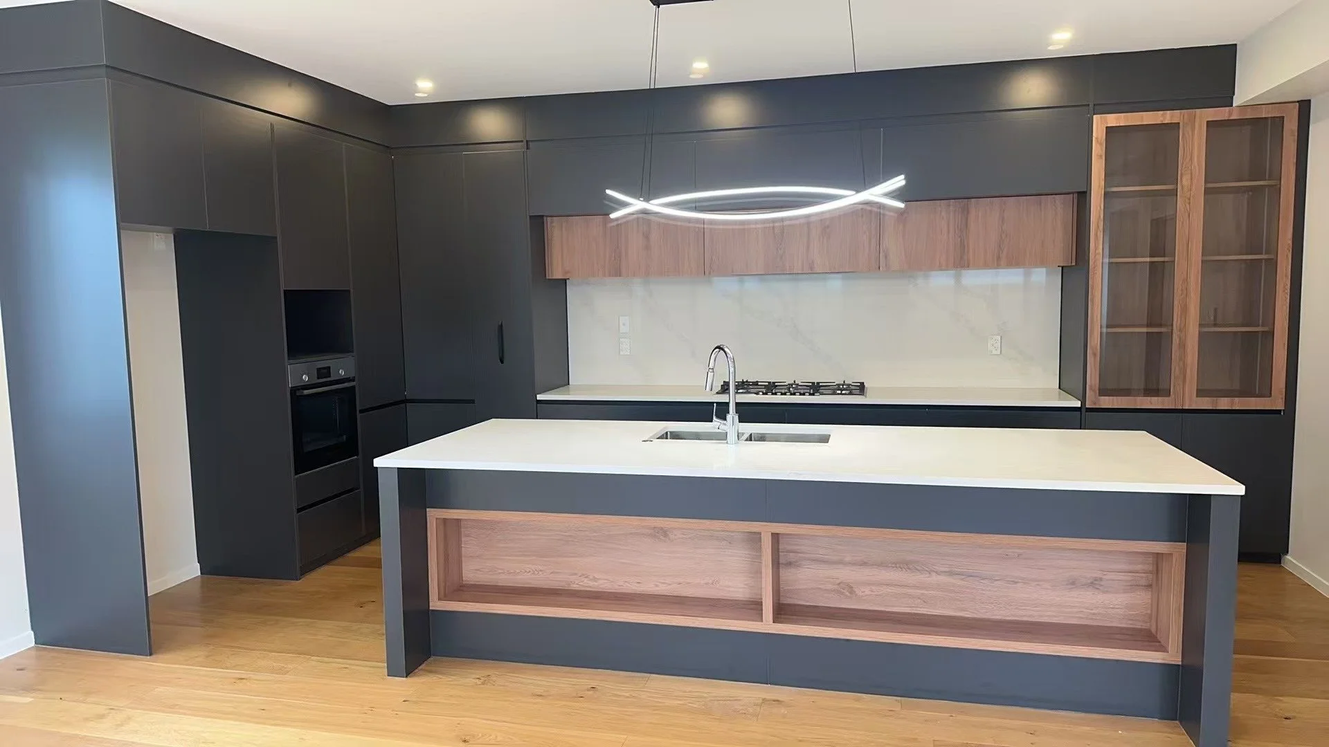 Modern kitchen with black and wood cabinetry, white island with sink, built-in oven, and a sleek mounted light fixture.