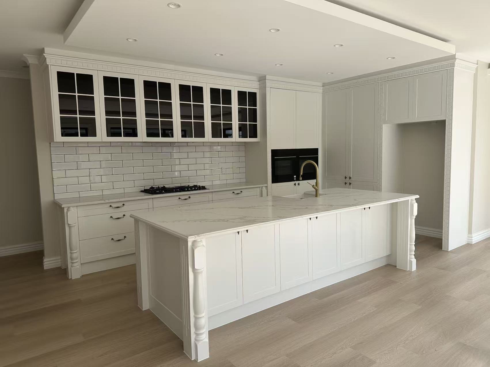 White kitchen with a large island, white cabinetry, a marble countertop, and glass-front upper cabinets, with appliance built-ins and hardwood flooring.
