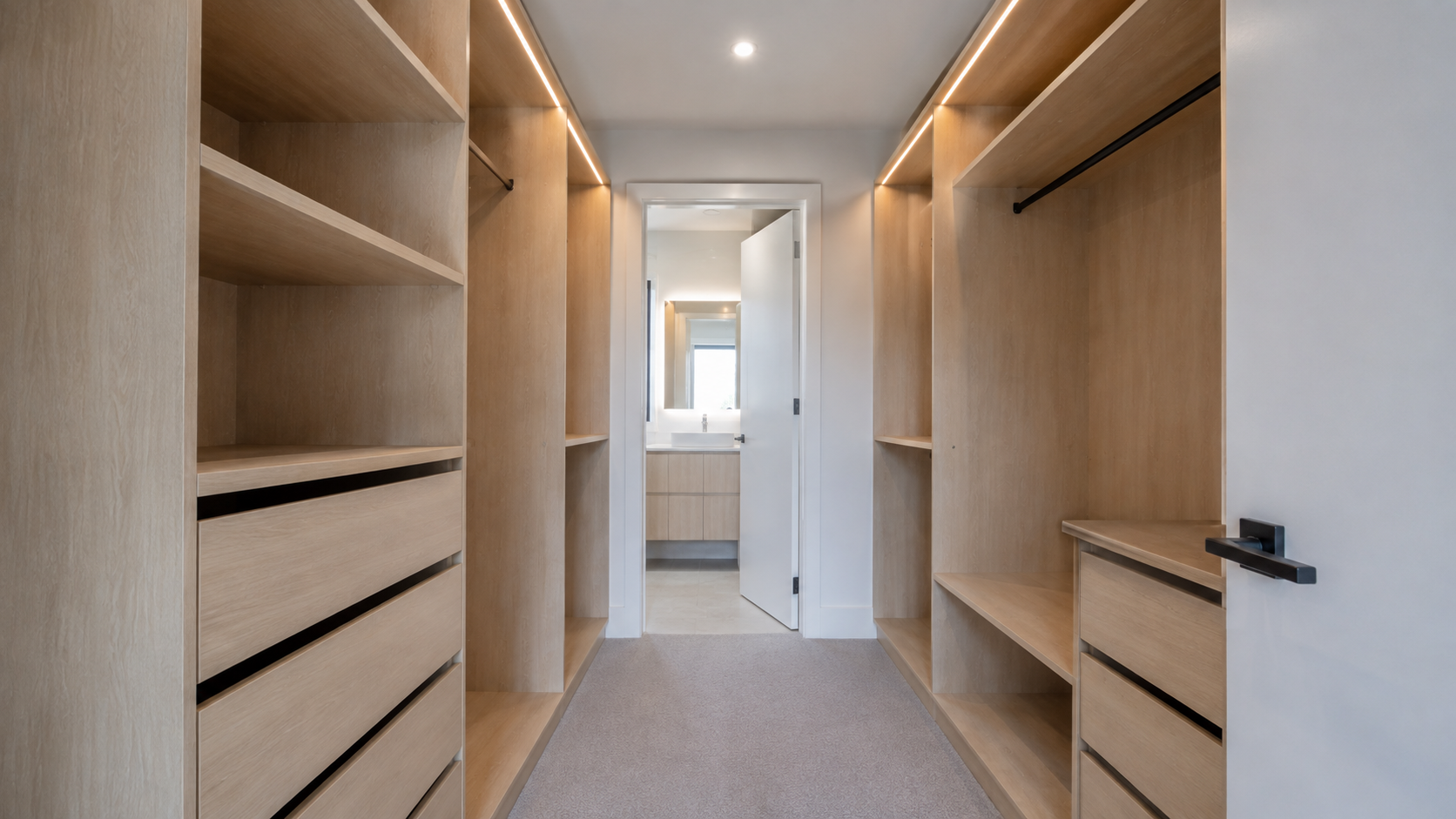 Empty walk-in closet with light wood shelving and drawers, black clothing rods, and a door leading to a bathroom with a vanity and mirror
