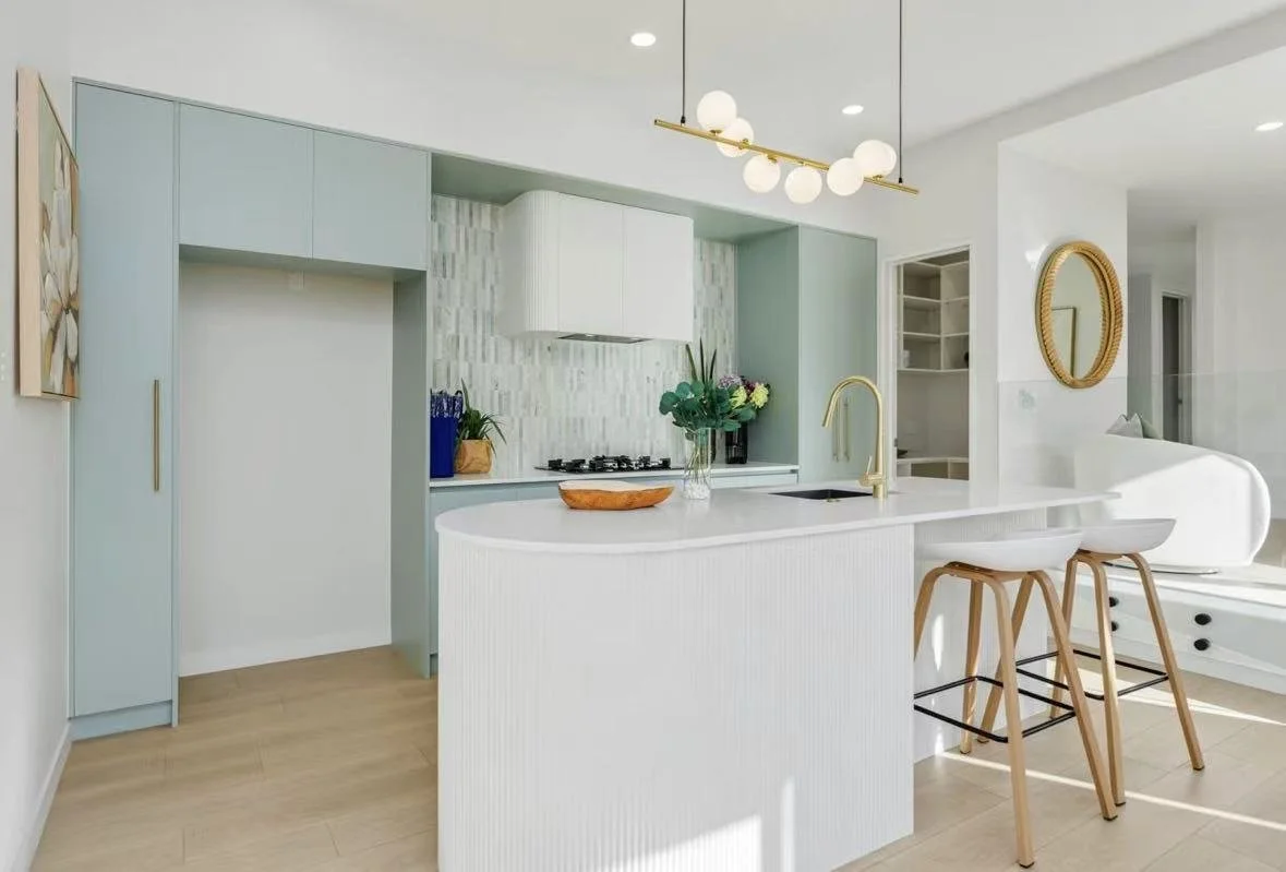 Modern kitchen with light blue cabinets, white countertops, gold fixtures, and a breakfast bar with two wooden barstools. Decor includes a vase with flowers and artwork on the wall.