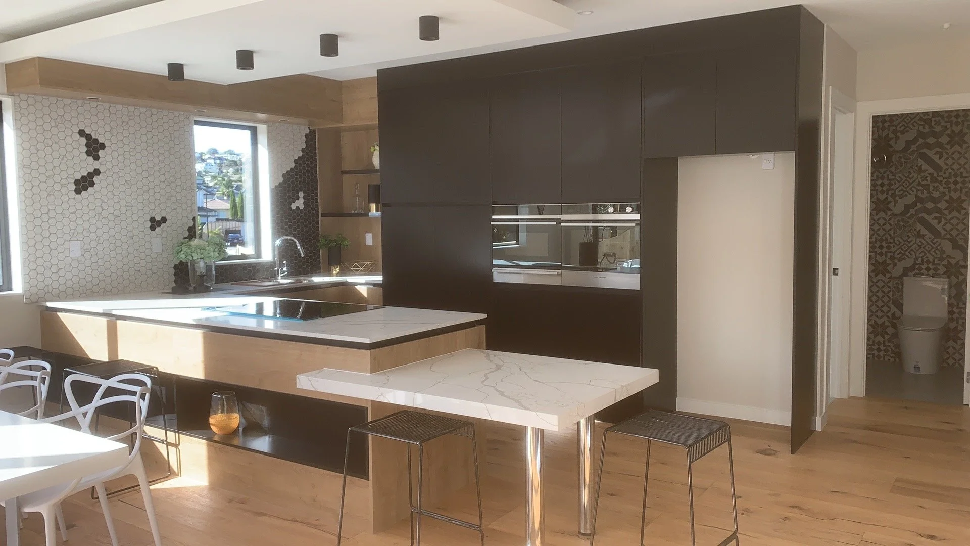 Modern kitchen with dark cabinetry, a central island with a white marble countertop, a breakfast bar, and a patterned backsplash. There's a view of a neighborhood through the window, and a bathroom with patterned wall tiles and a toilet is visible in