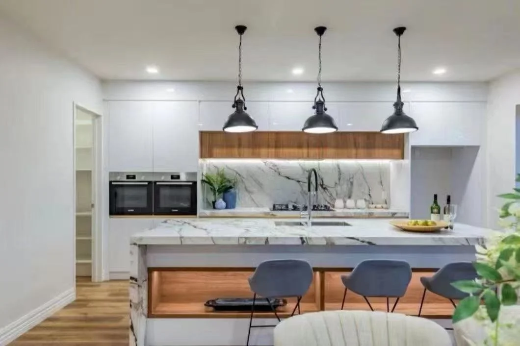Modern kitchen with white cabinets, marble countertops, three black pendant lights above the island, and wood accents. There are two built-in ovens, a plant in a blue pot, and three gray bar stools.