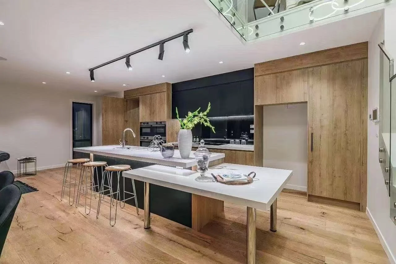 Modern kitchen with wooden cabinets, a white island with a sink, and bar stools. There is a large plant in a white vase on the island and some decorative jars. Recessed and track lighting illuminate the space, which features a glass staircase railing