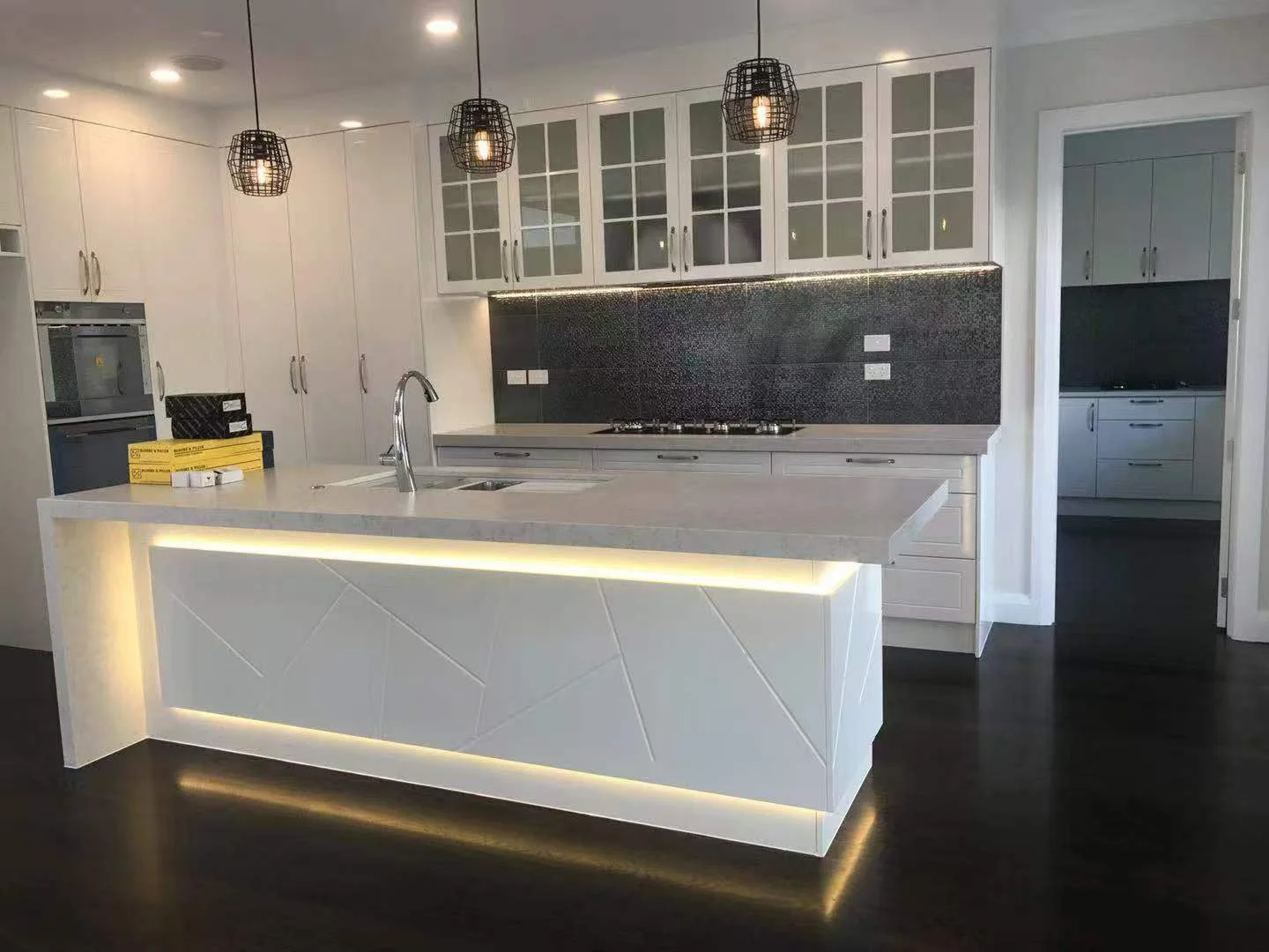 Modern kitchen with white cabinets, black backsplash, and a white island with LED lighting, pendant lights, and stainless steel fixtures.