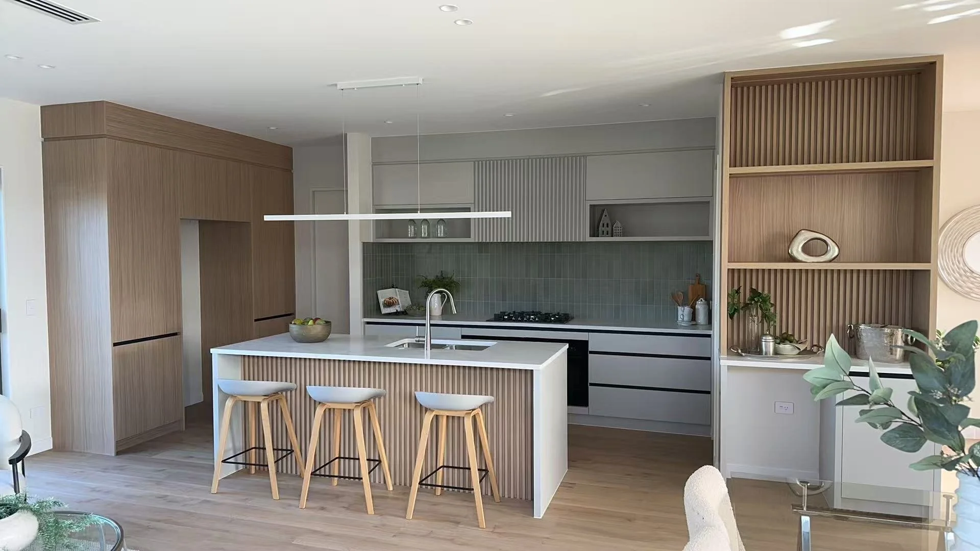 Modern kitchen with white island, wooden bar stools, gray cabinets, green tile backsplash, open shelving, and decorative accents.
