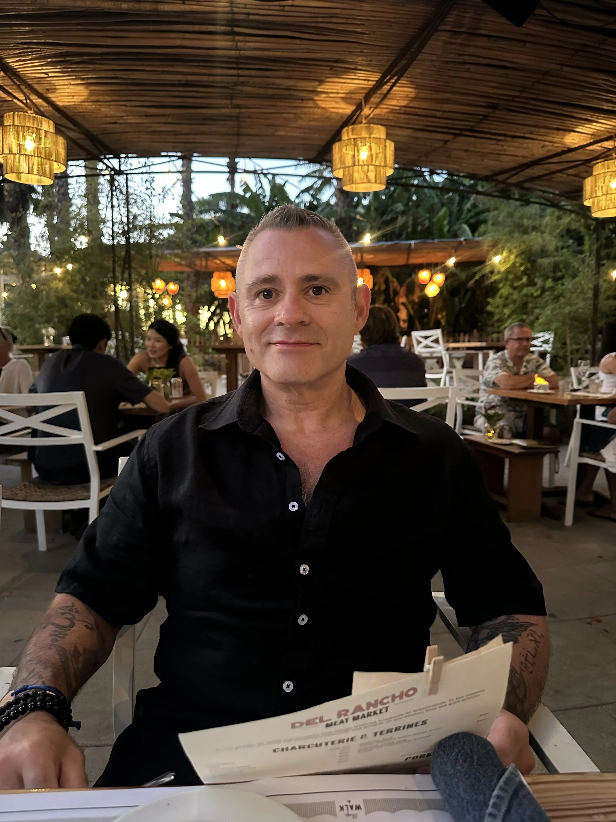 A man with tattoos on his arms, short hair, and wearing a black shirt, sitting at an outdoor restaurant with string lights and greenery, looking at the camera with a slight smile.