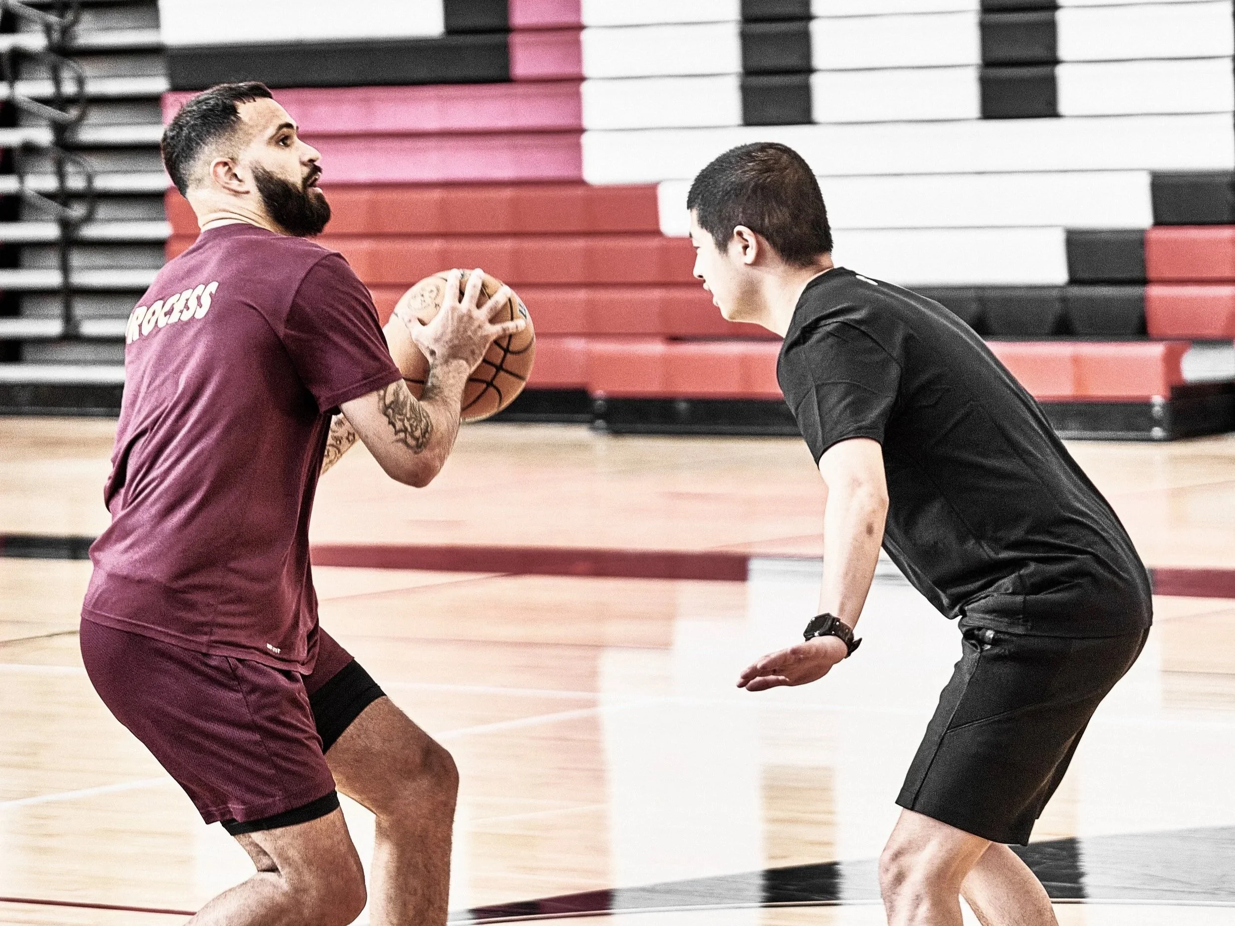 Two men practicing basketball in an indoor gym. One man with a beard and tattoos holding a basketball, and the other man in a black shirt and shorts squatting, facing each other. The gym has red, white, and black bleachers in the background.