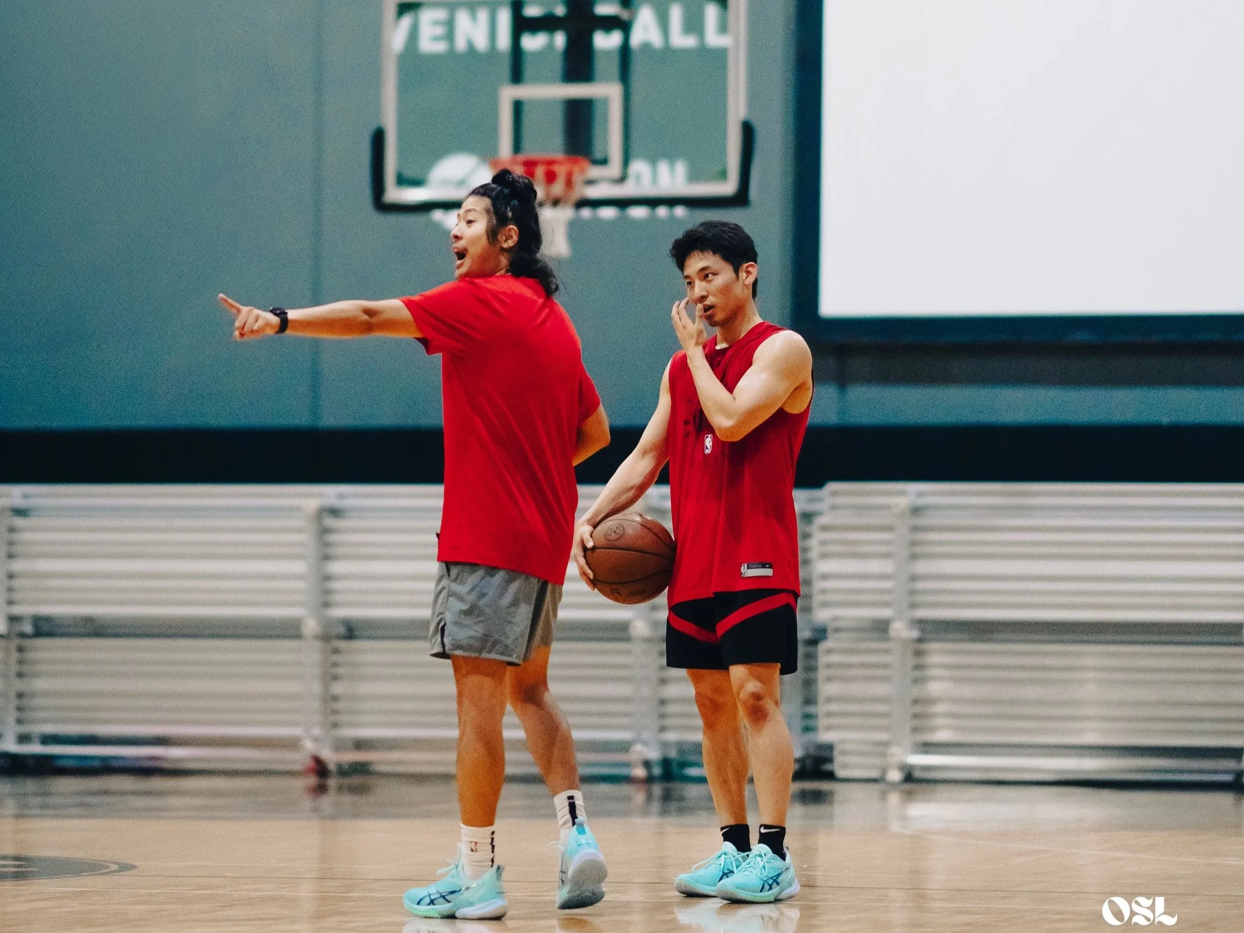 Two young men in red basketball jerseys on an indoor court, one holding a basketball, with a basketball hoop in the background.