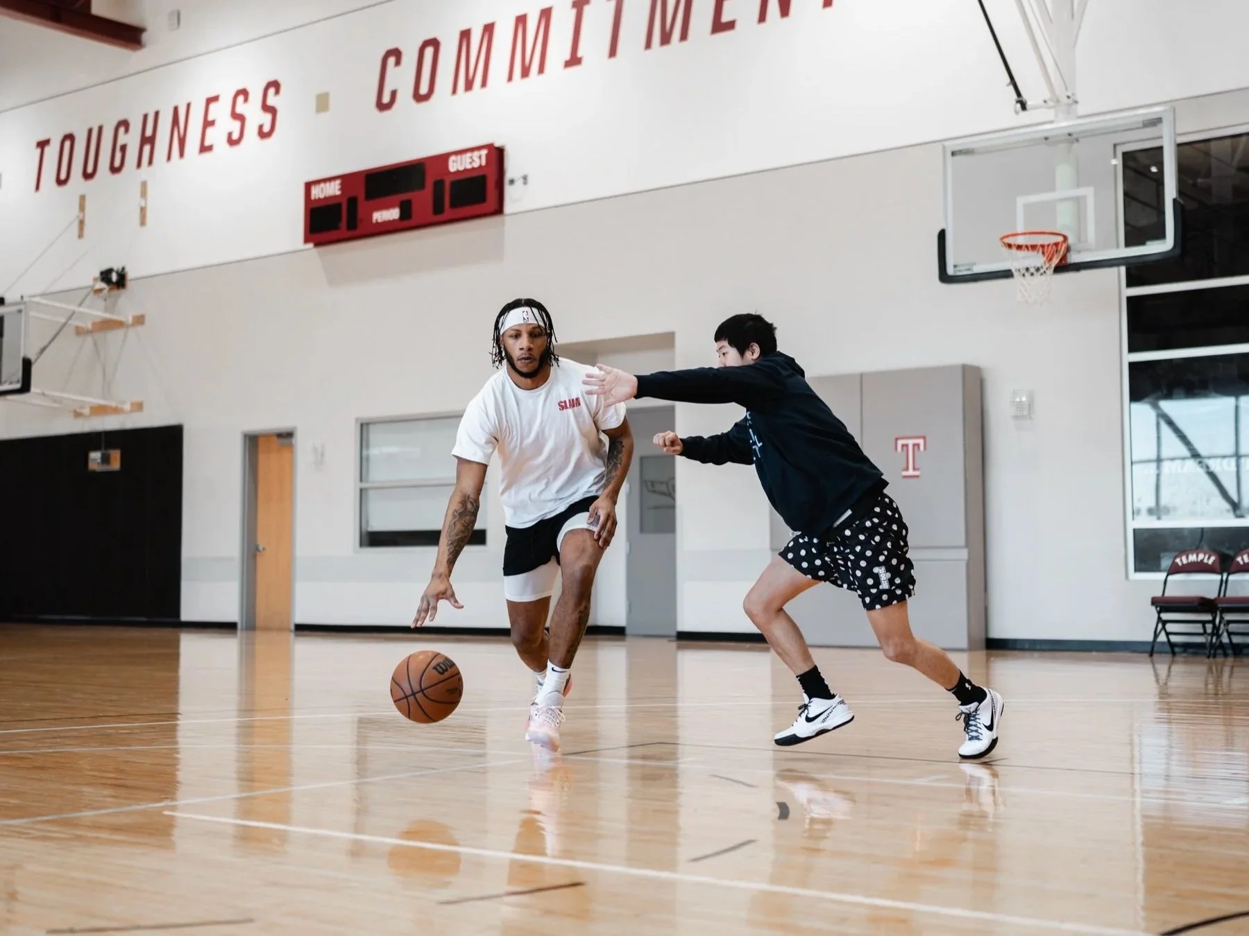 Two young men playing basketball on an indoor court, one dribbling the ball while the other defends. The court features a basketball hoop on the wall and a scoreboard above. The background includes a bench and a large wall with the words 'TOUGHNESS' and 'COMMITMENT' written prominently.