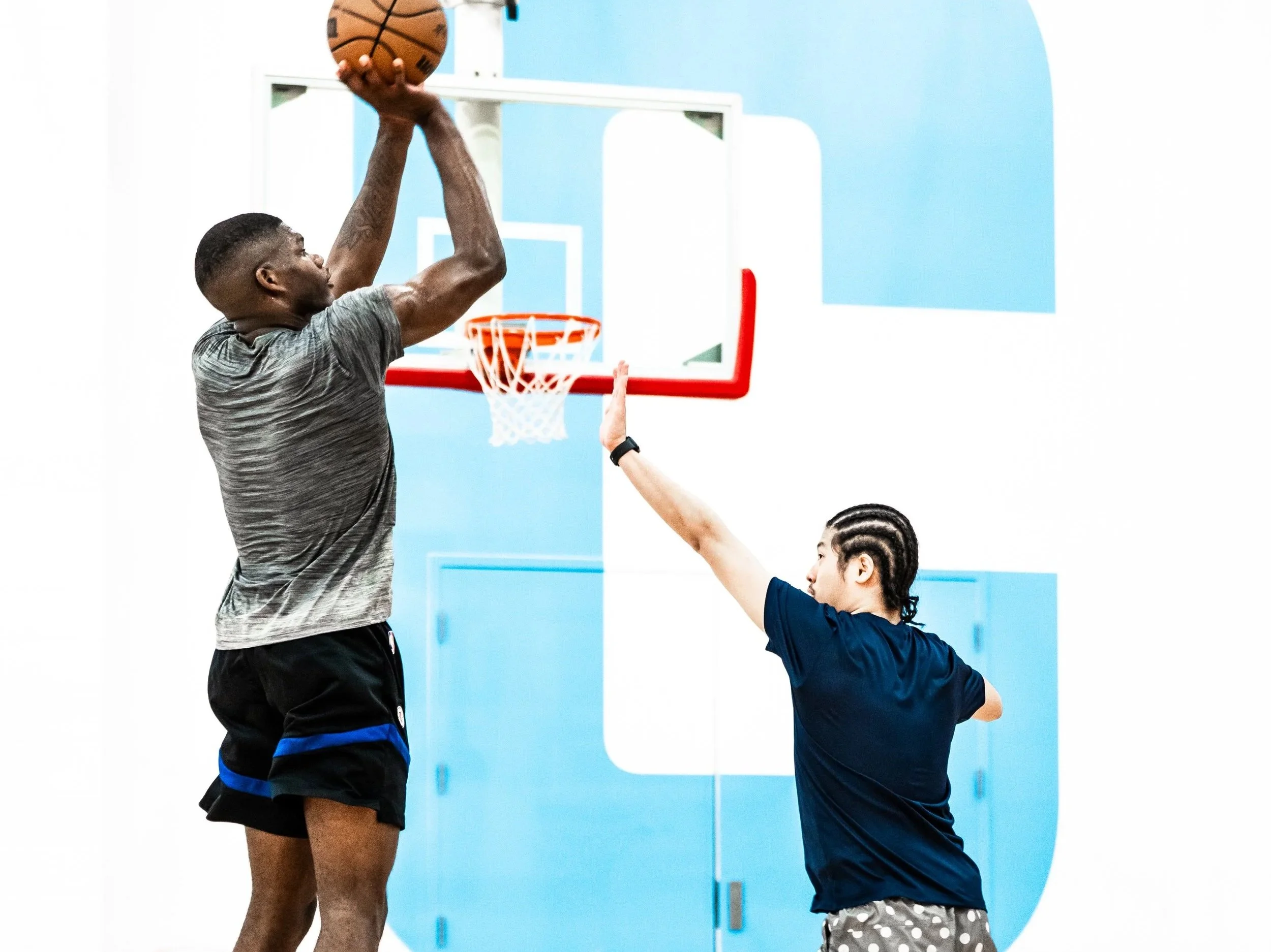 A man playing basketball is jumping to shoot a basketball while a girl is reaching up to block or catch the ball during a game in an indoor gym with a blue wall.