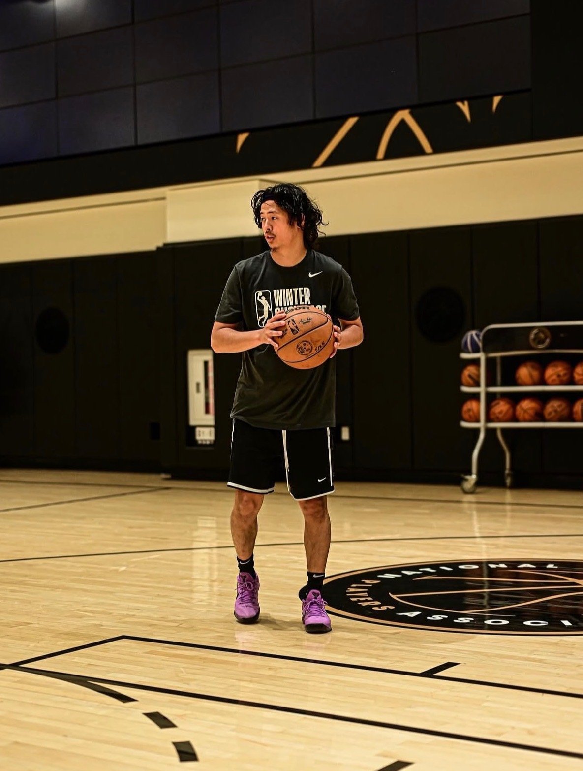 A man with long black hair, wearing a black t-shirt and shorts, is holding a basketball on a basketball court. There are multiple basketballs on a rack in the background.